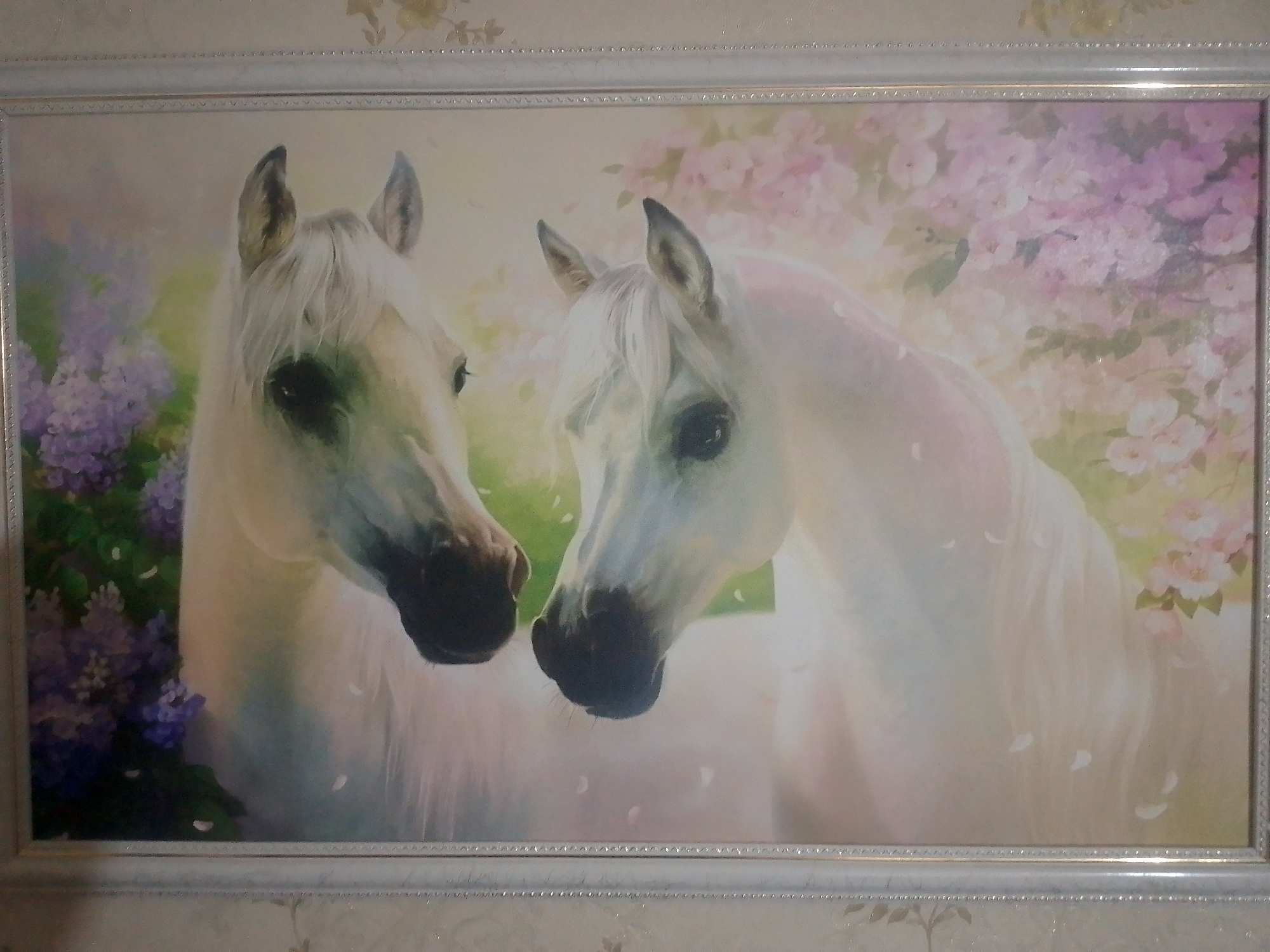 The image is a framed painting of two white horses with dark eyes and nostrils. The horses are positioned close together, facing the viewer. The left horse's head is slightly angled away from the viewer, while the right horse's head is angled slightly towards the viewer. They are set against a background of soft pink and purple flowers and greenery. The painting appears to be hanging on a wall with a patterned wallpaper. No specific location in Yessentuki, Russia, can be identified from the image alone.