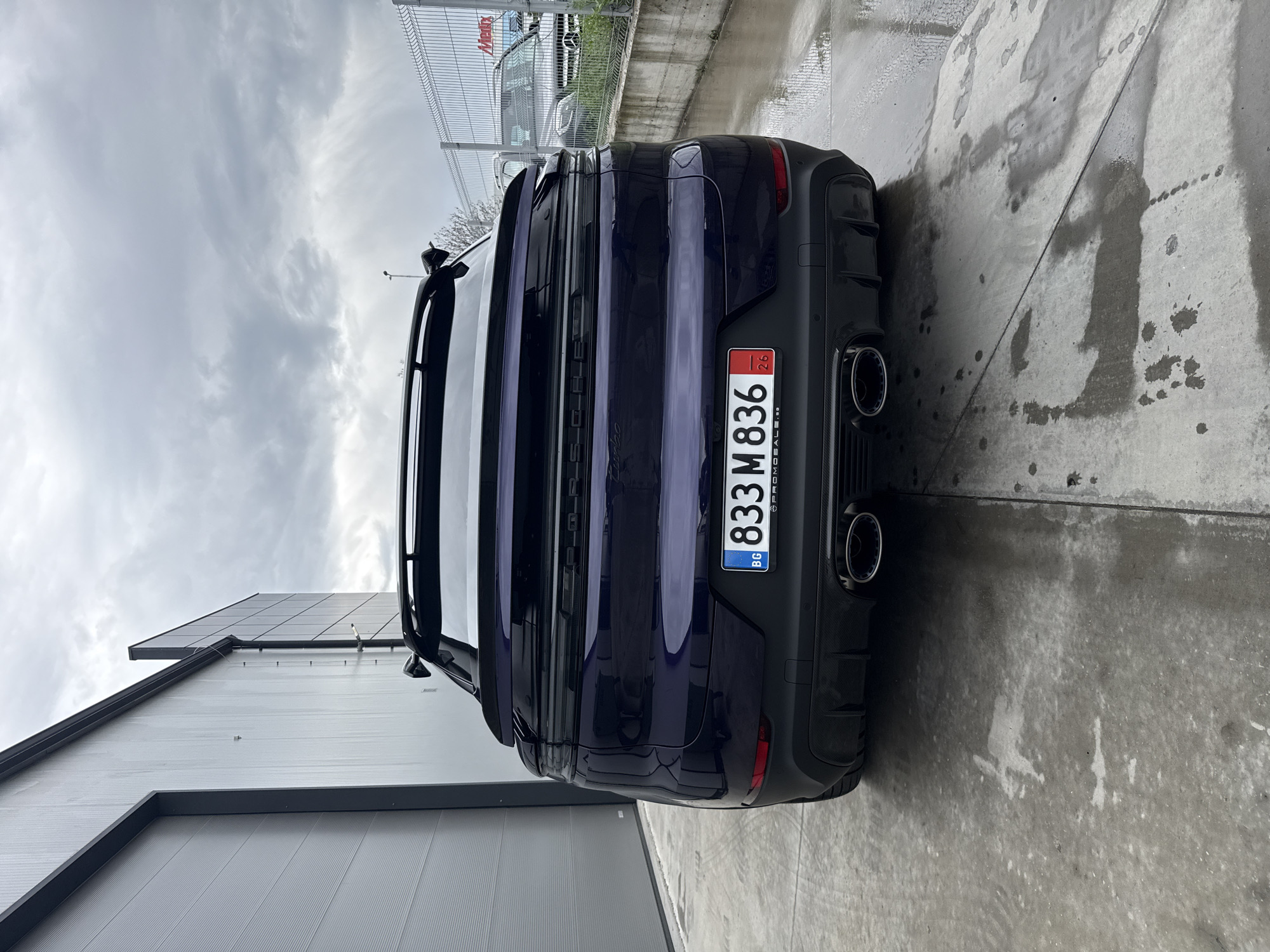 The image shows the rear of a dark blue Porsche Cayenne Turbo SUV, parked on a wet concrete surface, likely after rain. The vehicle has a Bulgarian license plate reading "BG 833 M 836". Behind the car is a tall metal fence with a sign visible on top, and a modern building with a light gray exterior wall and a dark gray roof. The sky is overcast. The location is plausibly an industrial or commercial area within Plovdiv, Bulgaria, judging by the appearance of the building and the license plate. No people are visible in the scene.