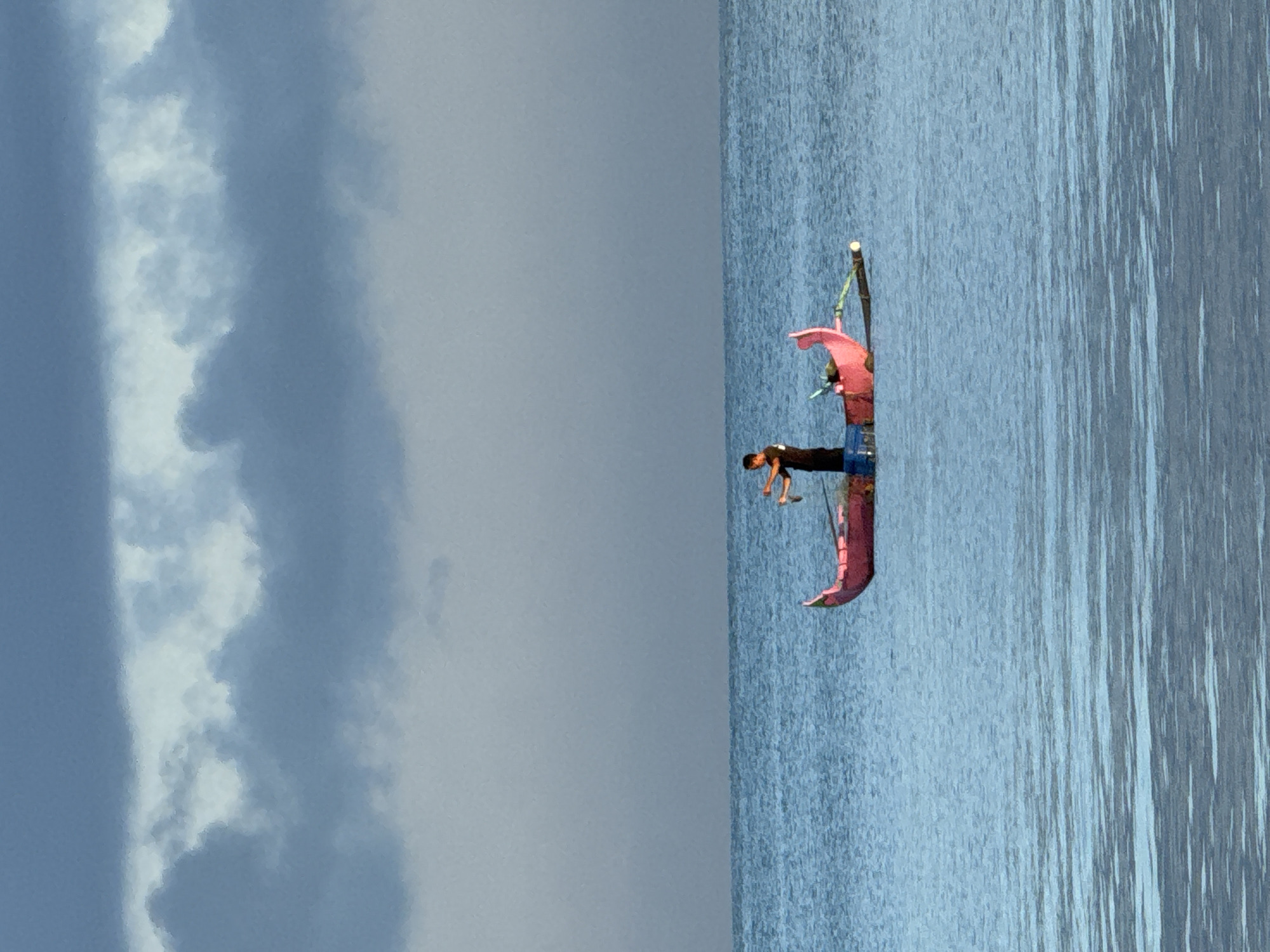 A man is standing in a narrow outrigger canoe in the ocean in Kuta, Indonesia. The canoe has a red outrigger arm and a light-colored bamboo support pole with a red decoration hanging from the end. The man is leaning over the edge of the canoe. The sky is cloudy and blue. The water is blue.