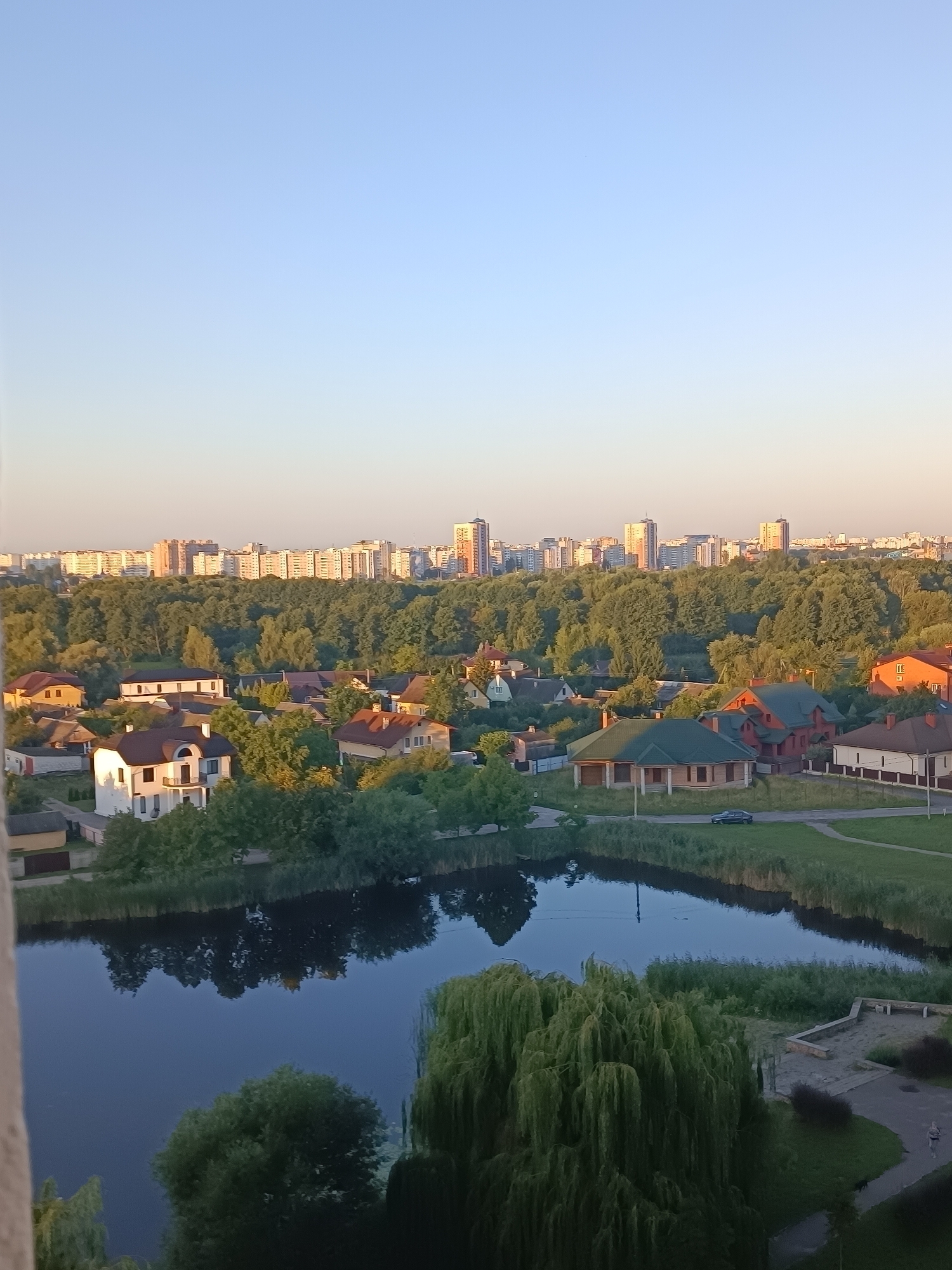 The image depicts a panoramic view of a suburban area with a lake in the foreground and a cityscape in the background. The foreground features a tranquil lake with reflections of trees and surrounding vegetation. A large weeping willow tree is prominently displayed on the right side of the lake. Beyond the lake, a cluster of houses, some with red tiled roofs, are nestled amongst lush green trees. In the mid-ground, a dense forest separates the suburban area from a distant cityscape. The cityscape is characterized by numerous apartment buildings and high-rise structures, indicating a more urbanized zone. The sky is clear and a pale blue, suggesting it is either early morning or late afternoon, with the sun casting a warm glow on the scene. A dark-colored car is visible on a paved road near the houses. The overall scene conveys a sense of peaceful coexistence between nature and urban development. While the image provides a scenic view, it does not contain sufficient identifiable landmarks or contextual cues to pinpoint a specific location within Brest, Belarus.