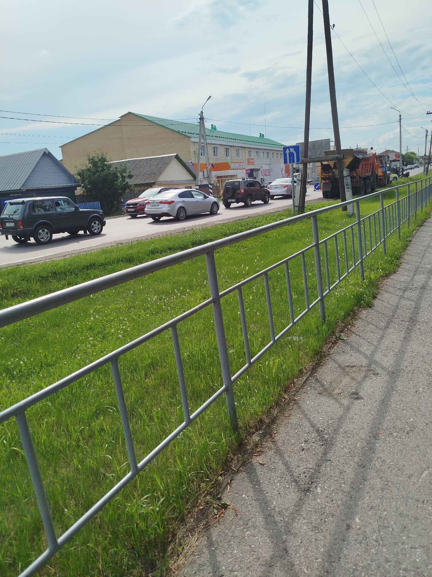 This image captures a scene on a street in Zavodoukovsk, Russia, with a focus on a road and its adjacent sidewalk. Several cars, including a dark green SUV, a red sedan, and a silver sedan, are visible on the asphalt road. Further down the road, construction vehicles, including a dump truck and a road roller, are present, suggesting roadwork is in progress. Buildings line the street, with one prominent building featuring a green roof and beige brick facade. A metal fence with vertical bars runs along the edge of the sidewalk, separating it from a grassy area. The sky is partly cloudy. There are no clearly identifiable individuals in the foreground.