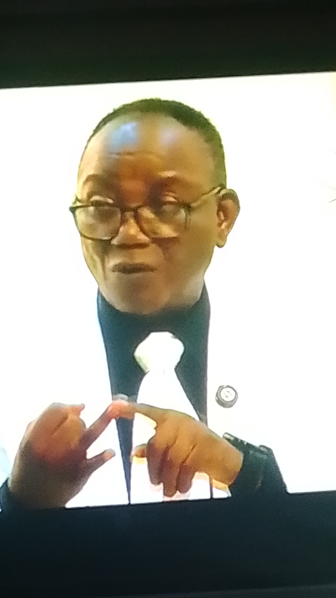 The image is a medium shot of a Black man wearing glasses and a white suit with a black collar and a white tie. He is facing forward and making a gesture with his hands, touching his index fingers together. The background is a plain white surface. The image quality is low, with a notable lack of detail and a slight tilt to the right. There is no visible indication of the location in Lokoja, Nigeria.