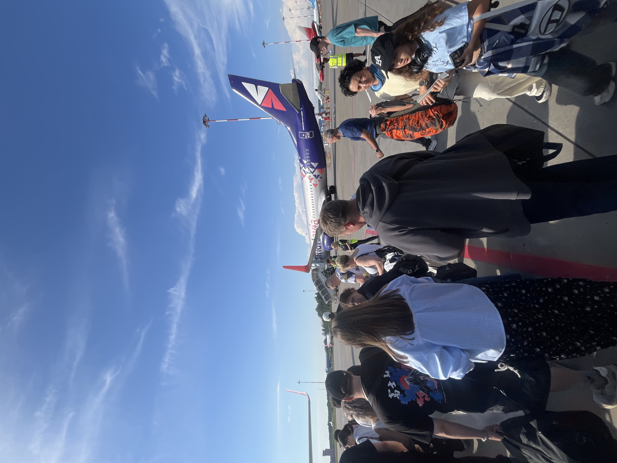 The image shows a group of people on an airport tarmac, boarding an airplane. The plane, with a purple and white livery, is parked on the tarmac, with a set of stairs leading up to the entrance. Several individuals are visible in the foreground and midground, some appearing to be passengers and others potentially ground crew. They are dressed in casual attire. The scene appears to be set during the daytime, with a clear blue sky and some scattered clouds. The lighting suggests it might be a sunny day. The presence of the airplane, tarmac, and boarding stairs clearly indicates an airport setting, and specifically, the act of boarding an aircraft. The people's postures suggest they are moving towards or waiting to board the plane. There's a sense of movement and anticipation associated with air travel. The overall atmosphere seems ordinary for an airport scene.
A visible text on the tail of the aircraft reads "VI" in large letters, and "RUS" is visible on the side of the fuselage. The registration number of the aircraft, "VP-RVT", is also visible on the fuselage.