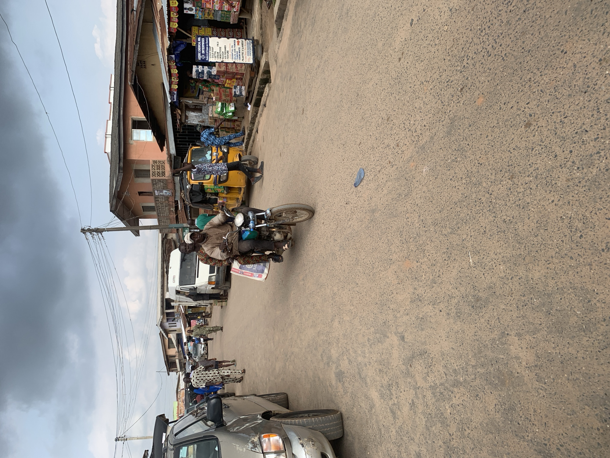 This media file depicts a street scene in Shagamu, Nigeria. The foreground features the front of a silver vehicle, partially obscuring the lower portion of the image. Dominating the mid-ground is a busy street with several people and vehicles. A yellow tricycle taxi is a prominent object, with a driver and at least one passenger. A motorcycle carrying two people is also visible, with one passenger holding a large bag that appears to be a cement sack.
Several other individuals are scattered throughout the scene, some appearing to be walking or standing near shops lining the street. The shops themselves are visible in the background, with displays of various goods and signage. One sign for "ADEMIC CHEMICAL NIG. ENTERPRISES" lists products like "LIQUID SOAP," "SOLID," "DETTOL," "CREAM," "INSECTICIDE," "BLEACH," and "AIR FRESHNER."
The weather appears overcast, with a mix of clouds in the sky. The time of day is difficult to ascertain precisely, but the lighting suggests it could be late morning or early afternoon. The overall impression is one of a bustling local marketplace or commercial area. The people are dressed in various attire, suggesting everyday life activities. There are no strong emotional cues visible. The ground is a paved road, showing signs of wear and tear. Power lines are strung across the scene, indicating the presence of infrastructure.