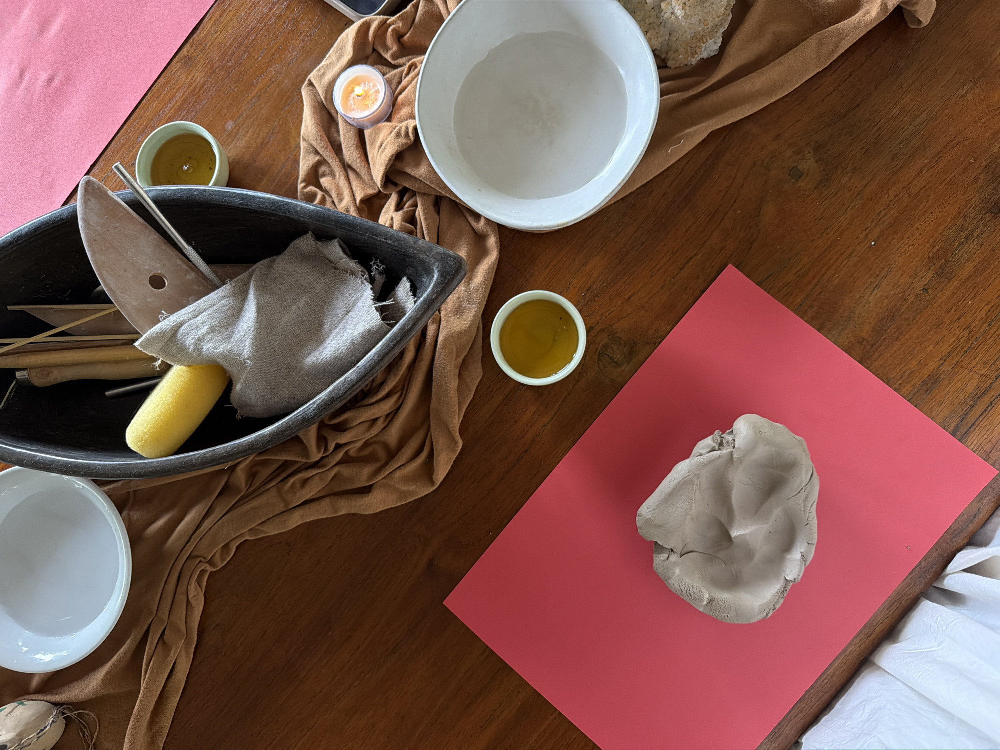 The image presents a top-down view of a rustic wooden table set up for a pottery or ceramics session. A large, dark rectangular bin occupies the mid-left, filled with various tools including wooden sculpting tools, a metal modeling tool, a yellow sponge, and a light brown cloth.
To the right of the bin, a large white bowl holds water, and a smaller white bowl is partially visible in the bottom left corner. A small, light green ceramic cup containing a yellow-orange liquid, possibly slip or oil, is positioned near the center. Another similar cup is seen in the top-left area. A lit candle in a clear glass votive holder casts a warm glow, positioned on a draped piece of crumpled, light brown fabric that extends across parts of the table.
In the bottom right, a rectangular red sheet or mat serves as a workspace, upon which rests a substantial lump of light gray clay, showing signs of having been worked or shaped. A piece of white material, possibly paper or cloth, is visible off the edge of the red mat in the bottom right corner. Another rough, light-colored material, perhaps unglazed ceramic or stone, is situated in the top right corner.
The overall scene suggests a calm and focused creative activity. The arrangement of tools and materials indicates preparation for hand-building or sculpting with clay. The setting appears to be indoors, possibly illuminated by natural light complemented by the candle's glow, creating a serene atmosphere. There is no visible text in the image. The activity takes place in Tegallalang, Indonesia.
