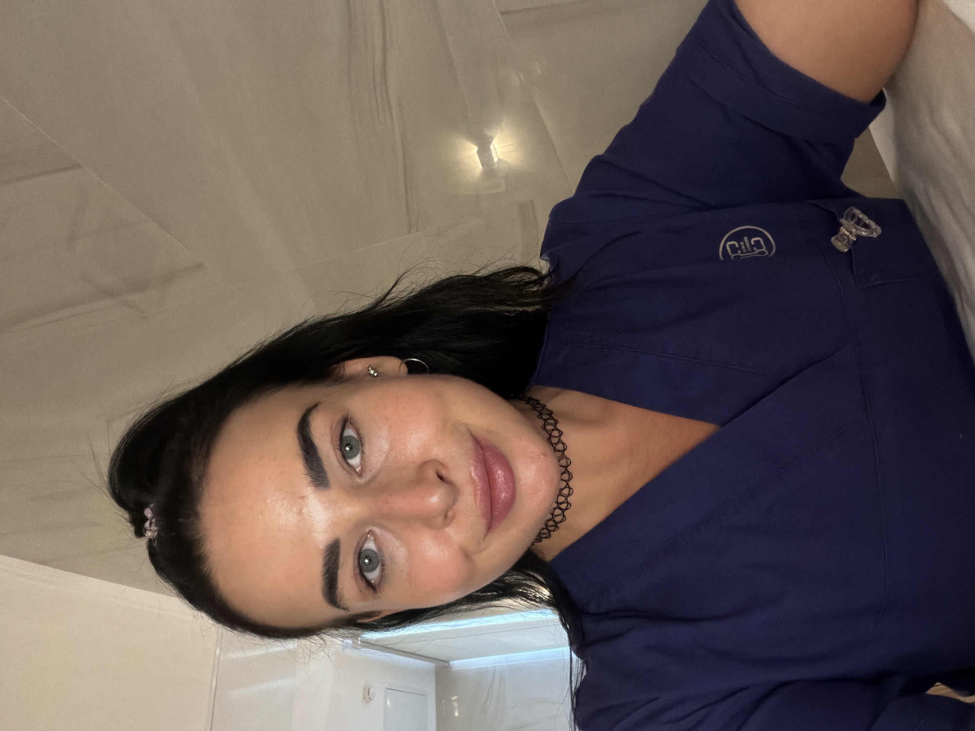 A woman with dark hair and gray eyes is posing indoors. She is wearing a dark blue V-neck scrub top with a logo on the left side and a bottle opener clipped to the pocket. She has a black choker necklace and earrings. The background shows a white ceiling with a light fixture and white curtains or fabric draped above. A small section of a white wall or door is visible in the lower left corner. There is no definitive indication of Saint Petersburg, Russia, in the image.