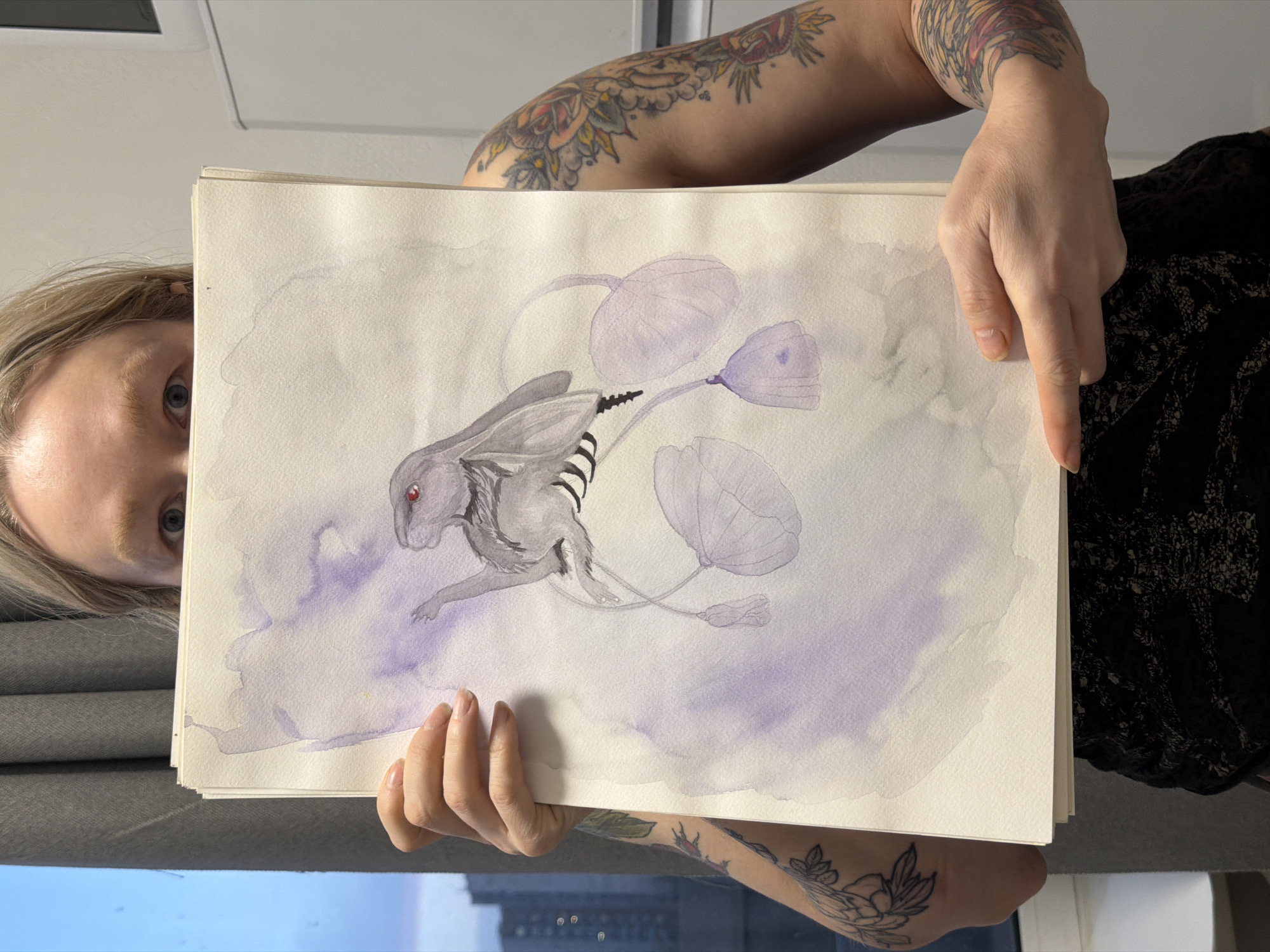 A person with light hair and tattoos is holding up a watercolor painting in front of them. The painting depicts a fantastical creature that resembles a rabbit with red eyes, black and white striped legs, and floating lavender-colored orbs attached by thin stems. The background of the painting is a wash of light purple watercolor. The person holding the painting has tattoos on both arms and is wearing a dark-colored top with a patterned design. A glimpse of their face is visible, with blue eyes looking at the camera. In the background, there is a window showing a cityscape, possibly an urban area with buildings. There is no evidence to confirm this image was taken in the city of Saint Petersburg, Russia.