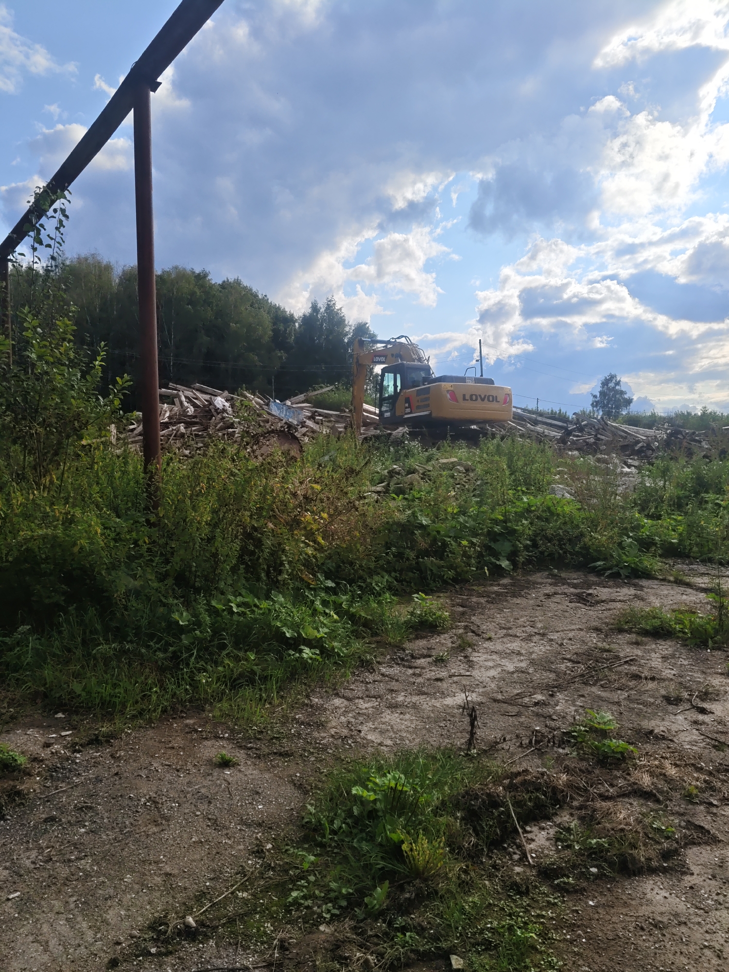 An outdoor scene in the village of Kukushtan, Russia, features a partially demolished structure and an excavator. A rusty metal beam, likely part of the original structure, stands vertically on the left, with overgrown vegetation surrounding its base. In the background, a pile of debris, consisting of wood, concrete, and possibly other building materials, is visible. A yellow excavator, identified by the "LOVOL" logo, is positioned atop the debris pile, suggesting demolition or clearing work is in progress. The surrounding environment is characterized by a mix of lush green vegetation and a dirt or gravel ground. The sky above is partly cloudy with a mix of blue sky and white cumulus clouds.