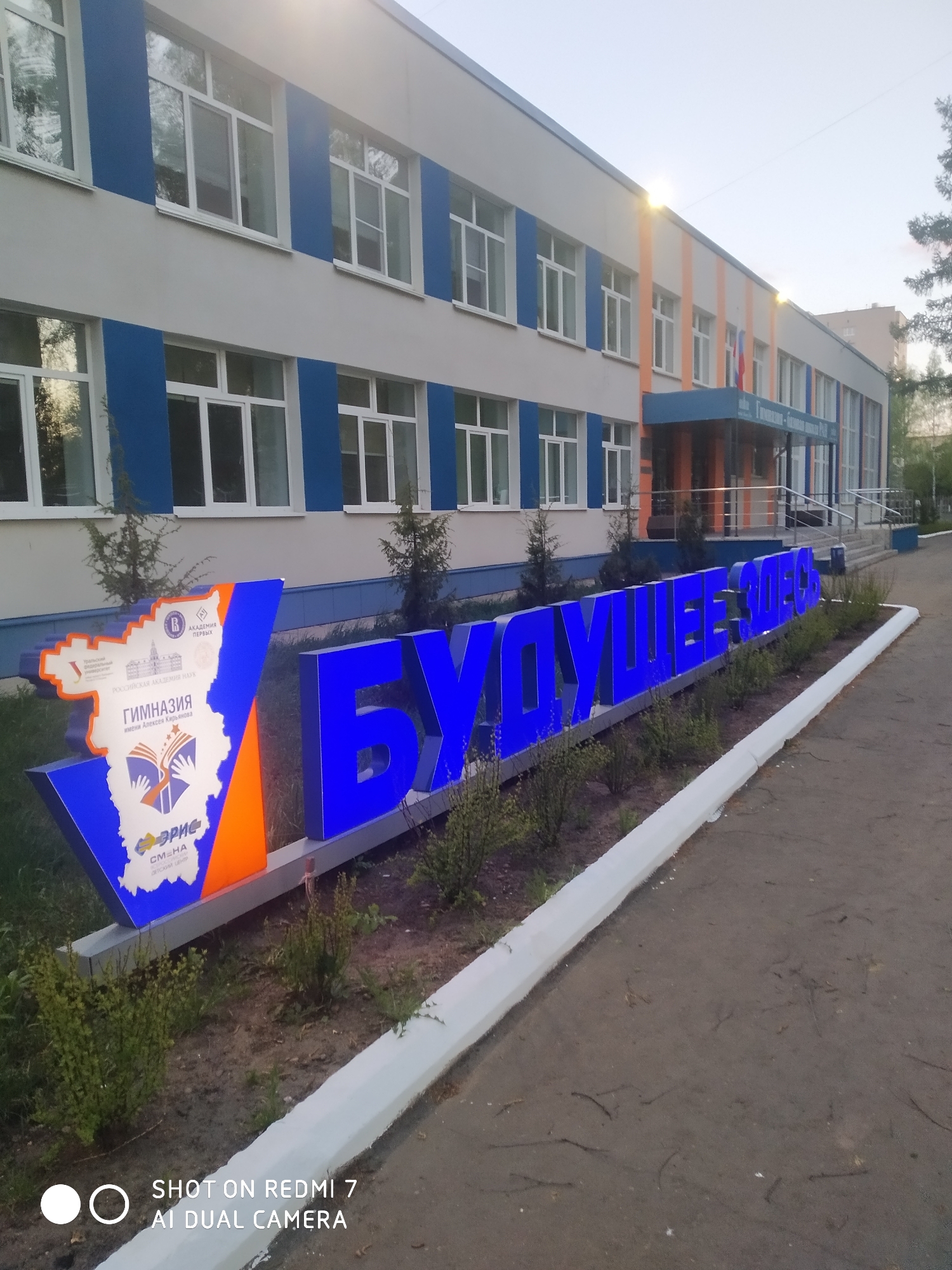 Here's a description of the image:
The image depicts a building with a sign in front of it, likely located in the city of Chaikovsky, Russia. The building has a multi-story structure with a facade featuring a pattern of alternating gray, blue and orange panels around the windows. It has multiple white-framed windows.
In the foreground, there is a large, illuminated sign. The sign contains an emblem with images related to education and academic institutions. A blue word is illuminated and displayed prominently. There are small plants and bushes around the sign. A gray pavement is in the foreground with a white curb. The text at the bottom indicates that the photo was taken with a Redmi 7 AI Dual Camera.