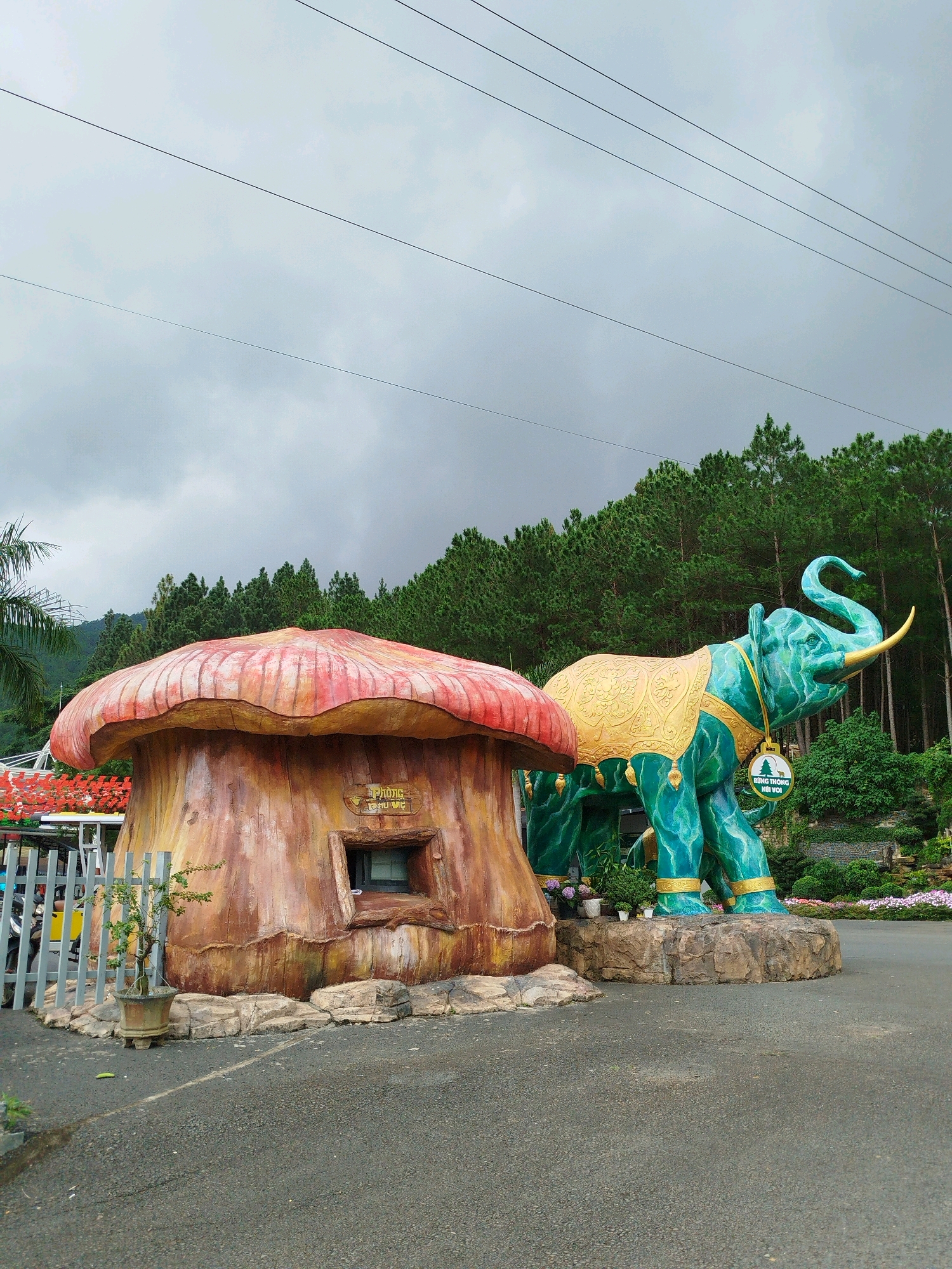 In K'Long, Vietnam, a paved area features a large, stylized sculpture of a green elephant adorned with a gold cloak and collar. The elephant is positioned next to a mushroom-shaped building with a red and yellow cap and a brown, textured base. The mushroom structure has a small, dark window. Behind the sculptures, a dense forest of pine trees lines a hillside under a cloudy sky. Power lines are visible stretching across the upper portion of the image.