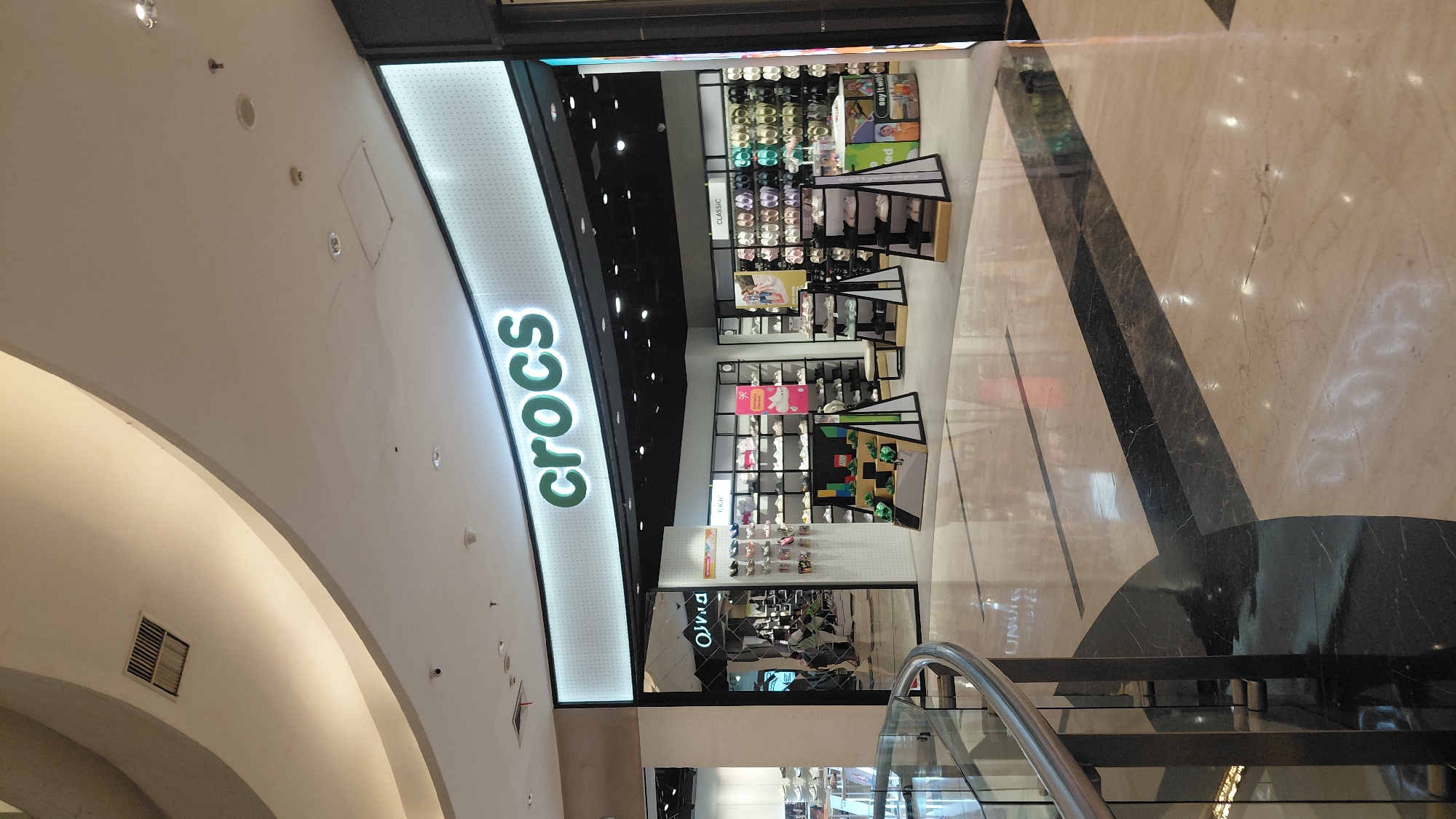 The image shows the exterior of a Crocs store in a shopping mall. The store has a large, illuminated "crocs" logo above the entrance. Inside the store, shelves are stocked with various styles of Crocs footwear. Several display stands are positioned in front of the store, showcasing different shoe models. The mall's flooring is made of polished marble with a geometric pattern. The perspective is from a slightly elevated viewpoint, looking towards the store entrance from a walkway. Based on the available information, this Crocs store is located in the city of Purwokerto, Indonesia.