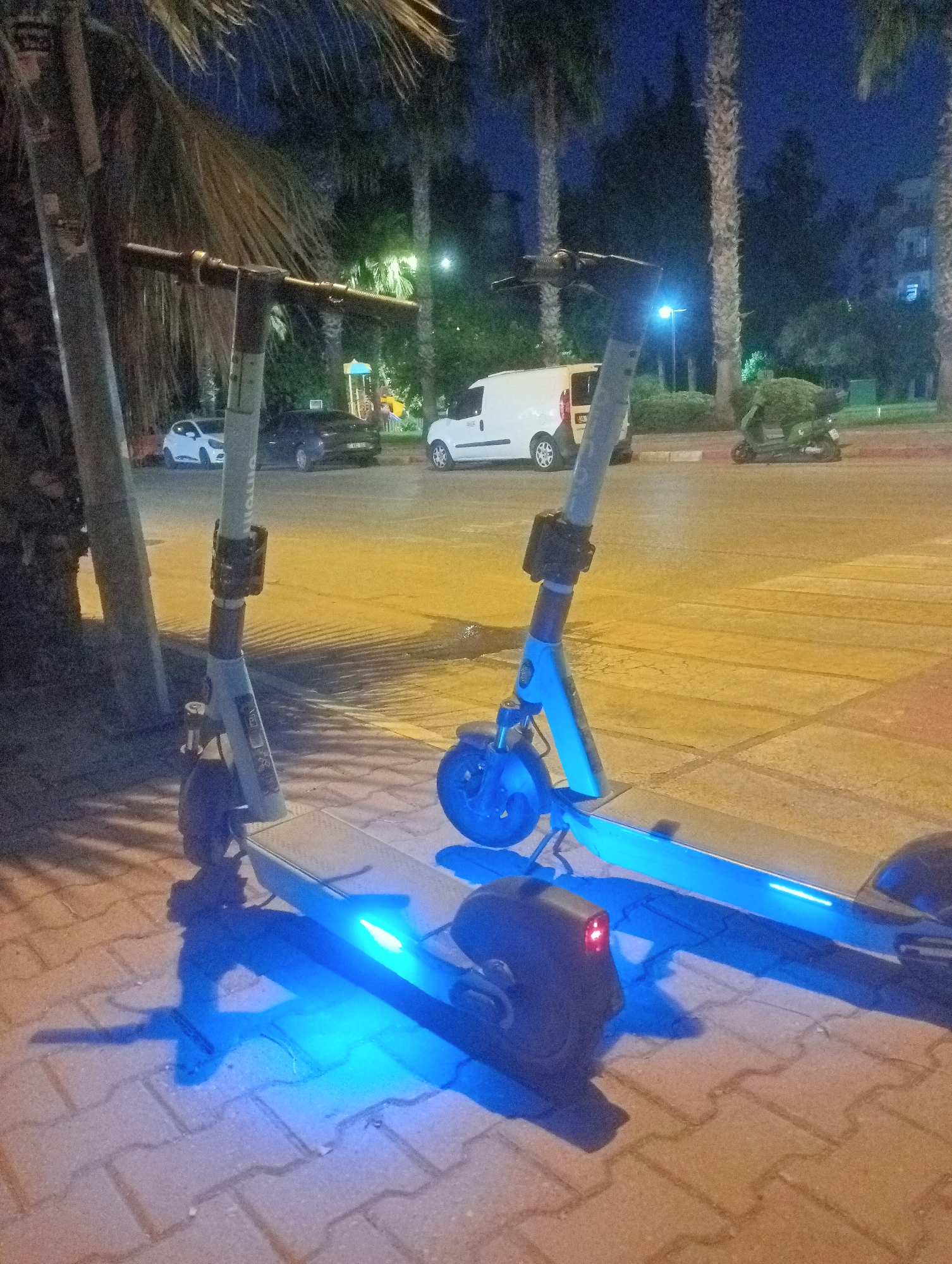 In Antalya, Turkey, on a street at night, two electric scooters with blue LED lights are parked on a sidewalk. The scooter in the foreground is facing right, while the second scooter is slightly behind and to the right. Palm trees and other foliage are visible in the background, along with a white van, a dark sedan, a white car, and a scooter on a distant street. Buildings and a lamppost are also present. The ground is paved with interlocking bricks.