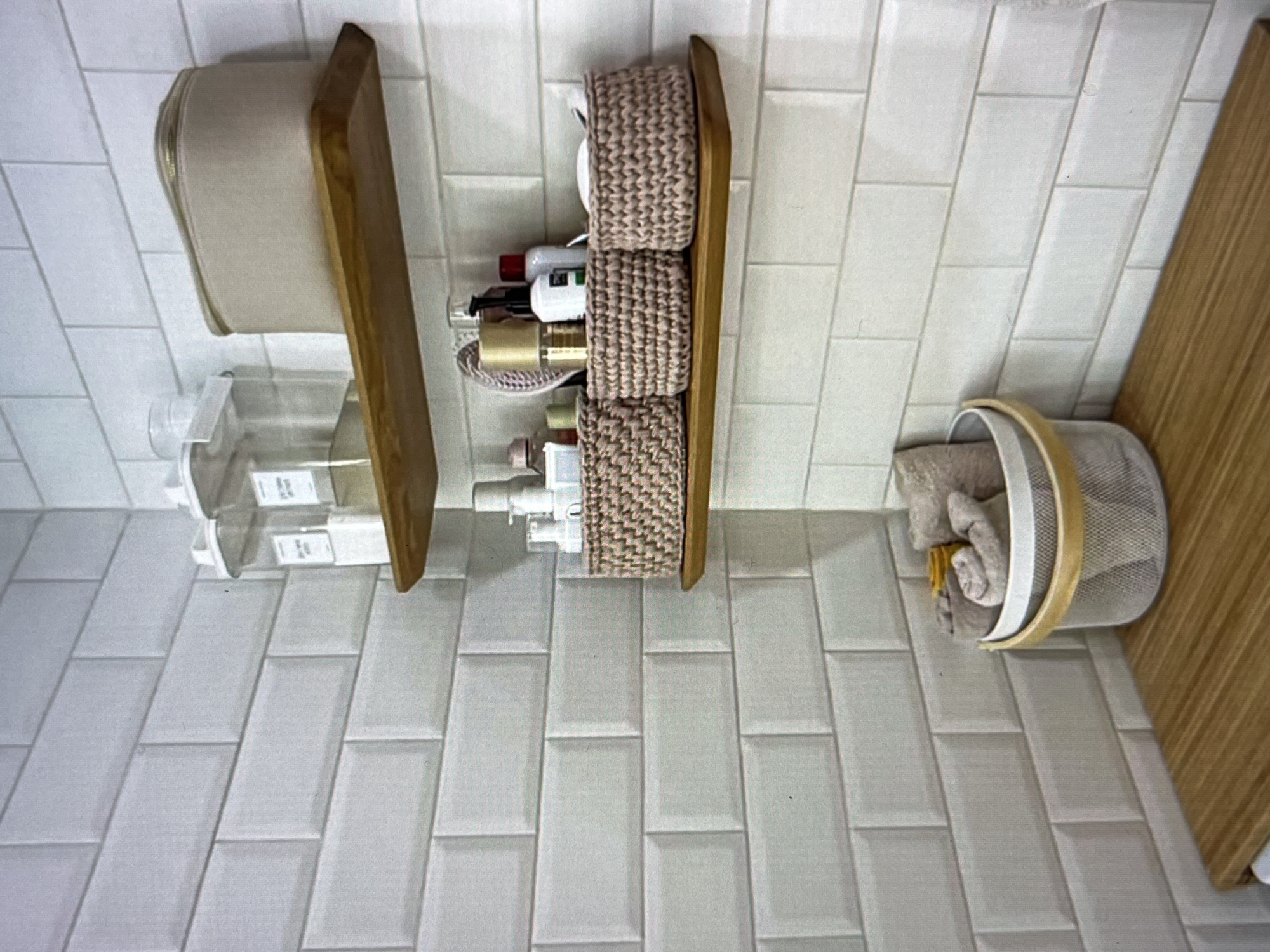 The image shows a corner of a bathroom with white subway tiles on the walls. There are two wooden shelves mounted on the tiled wall. The top shelf holds a beige zippered cosmetic bag and two clear plastic containers with white lids. The bottom shelf displays three small crocheted baskets, two of which contain toiletries or makeup. The third basket, a larger mesh one with a wooden handle, sits on a wooden countertop and contains rolled-up towels and a small yellow item, possibly a loofah. The overall scene suggests a tidy and organized bathroom space. There are no people or actions depicted in the image. The location in Dubai, United Arab Emirates is not discernible from the visual evidence.