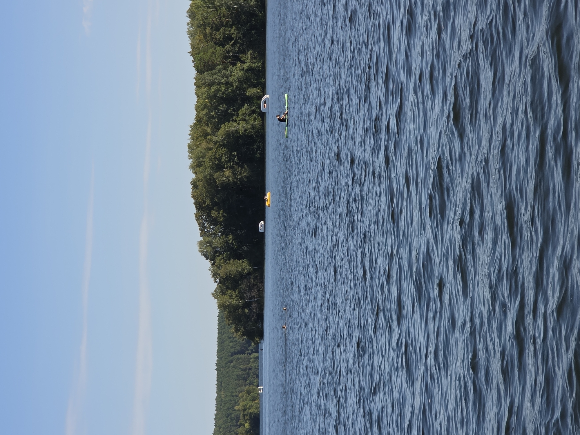 The image shows a wide, blue lake with moderate waves, under a clear sky with thin, horizontal clouds. In the foreground, the water fills the entire frame, showing its rippled surface. In the mid-ground, there are several individuals and watercraft. A person is seated on a green paddleboard in the center-right of the frame. To the left of the paddleboarder, two individuals are swimming in the water. Further in the distance, there are two white pedal boats, one on the left and one on the right. On the far left, a small white structure is visible near the shore. The background features a dense forest of green trees lining the lake's edge, with a strip of lighter green trees and a darker, pine-forested hill beyond. The scene appears to be a recreational area on a lake, likely in Chebarkul, Russia, given the mention of the city and the commonality of lakes and forests in the region.