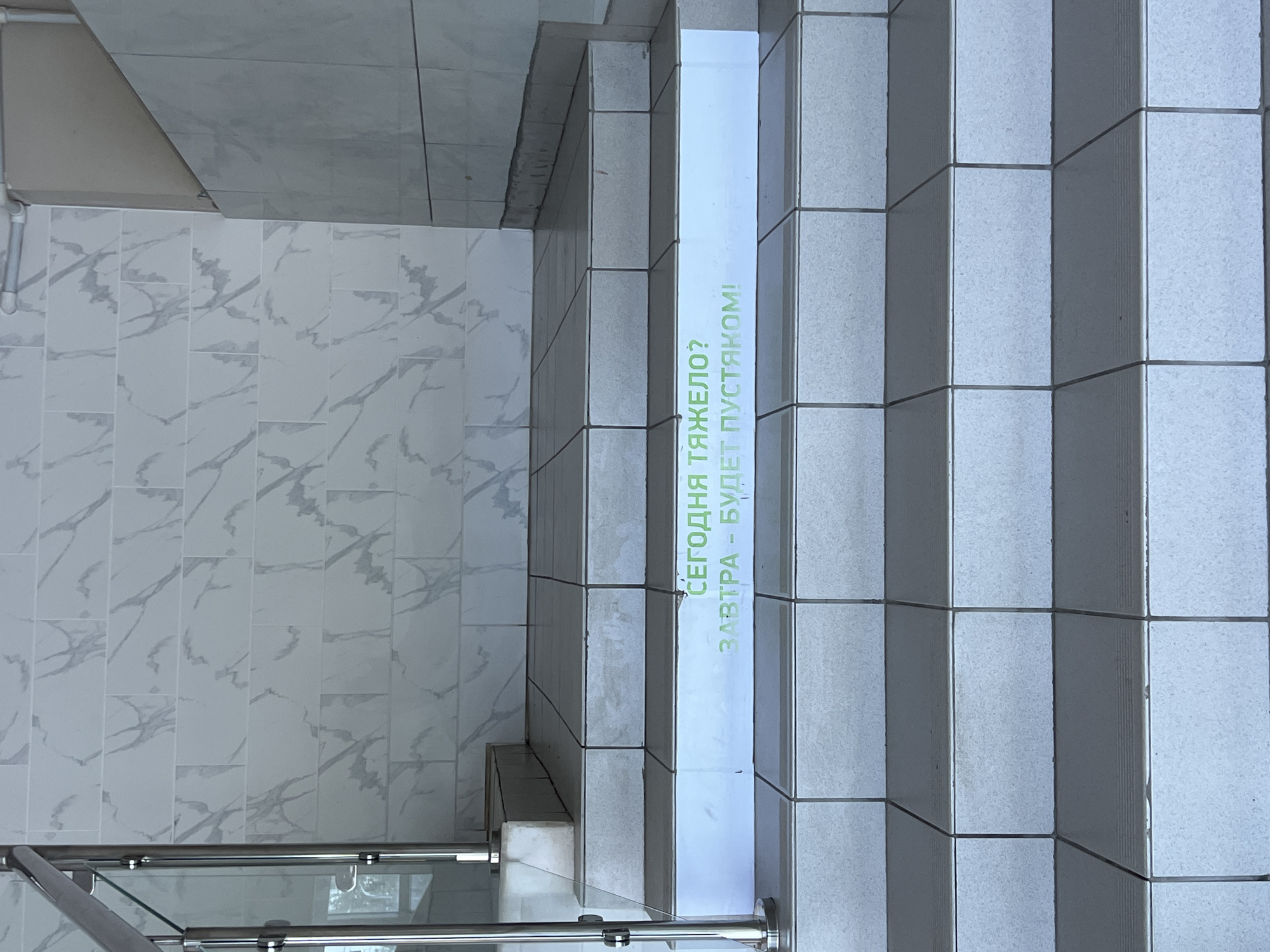 The image shows a staircase in what appears to be an indoor location. The stairs are covered in light gray tiles. A vertical sign is placed on the side of the stairs with green text in Russian that reads "СЕГОДНЯ ТЯЖЕЛО? ЗАВТРА - БУДЕТ ПУСТЯКОМ!", which translates to "Is it hard today? Tomorrow it will be easy!". A metal railing with glass panels is visible at the bottom of the stairs. The floor to the left of the stairs is tiled with white marble-patterned tiles.
Given the text is in Russian, it is likely that this image was taken in a Russian-speaking location. Without additional information, it is not possible to confirm if it is located in the city of Chaikovsky, Russia.