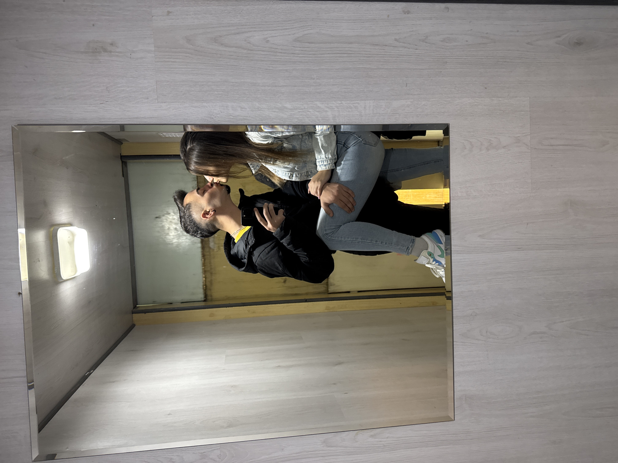 A mirrored reflection depicts a couple kissing in what appears to be a small elevator or confined space. The male figure has short, dark hair and wears a black jacket with a yellow collar, along with blue jeans and sneakers. The female figure wears a light denim jacket and blue jeans. They are embracing. The background suggests the inside of an elevator, with light-colored walls and a light fixture visible. The reflection is set against a backdrop of what looks like a light-colored, wood-patterned floor. The location in Sofia, Bulgaria, is not possible to determine from the image.