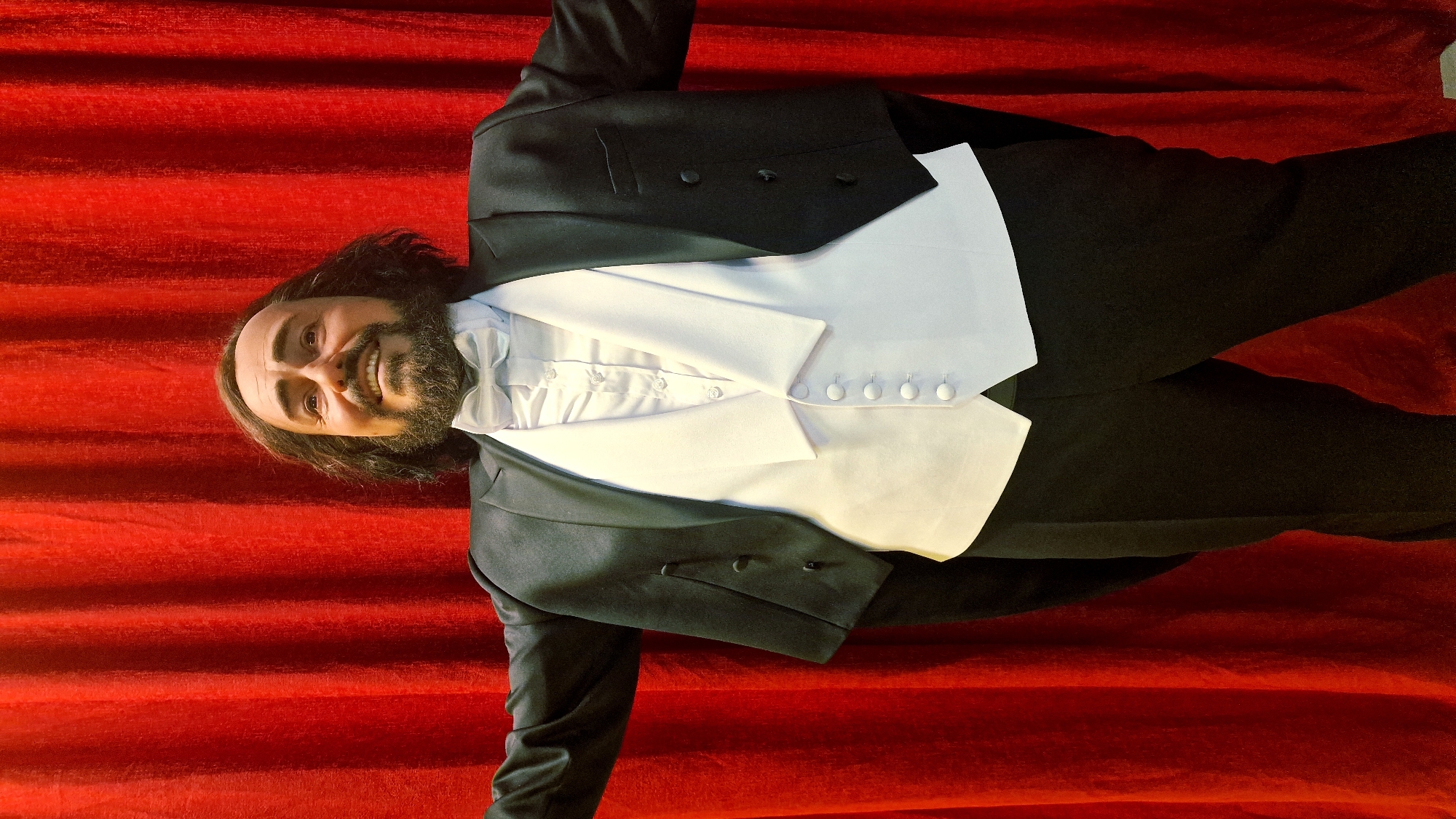 A man is depicted from the waist up, smiling warmly and looking directly at the viewer. He has long, dark, wavy hair and a full dark beard and mustache. His complexion is fair, and his eyes are dark. He is dressed in formal attire, including a black tuxedo jacket with satin lapels, a crisp white pleated dress shirt, a white waistcoat with five prominent white buttons, and a matching white bow tie.
The background consists of a rich, vibrant red fabric, likely a plush curtain, displaying horizontal folds and a soft texture. This setting suggests an indoor location such as a theater, concert hall, or a formal event. The man's expression conveys happiness, approachability, and confidence as he poses for the photograph. The lighting is even and highlights the details of his attire and facial features. There is no visible text in the image.