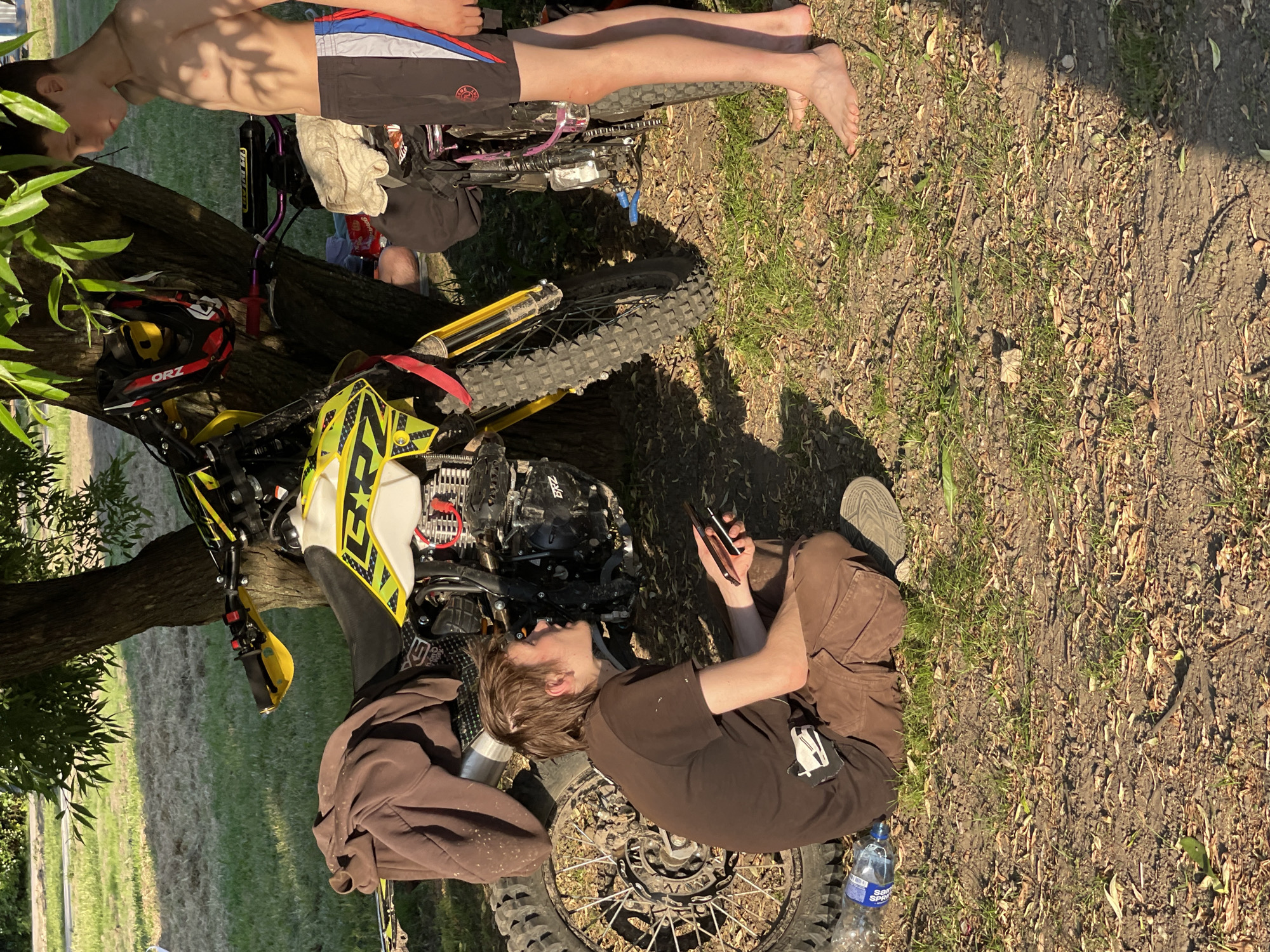 The scene, depicted in what is identified as the city of Moscow, Russia, features two young males and two dirt bikes resting beneath a deciduous tree. The ground consists of dry soil mixed with sparse green grass and scattered light-colored debris. Strong, natural sunlight casts distinct shadows, indicating either morning or late afternoon.
On the left, a male, appearing to be in his late teens or early twenties, sits cross-legged on the ground. He has light brown, somewhat disheveled hair and fair skin. He wears a dark brown short-sleeved t-shirt with a white graphic on the lower left side, brown cargo pants, and a dark-colored athletic shoe visible on his right foot. He holds two black smartphones in his left hand and gazes upwards and to his right.
To his right, a second male, appearing to be a pre-teen or young teenager (approximately 10-14 years old), stands barefoot. He has short, dark hair, fair skin, and a lean physique. He is bare-chested and wears black athletic shorts with vertical red, white, and blue stripes on the outer sides. He looks downwards and slightly to his left.
Between the two individuals and leaning against the tree trunk is a prominent yellow and black dirt bike, branded "BRZ" on its engine casing and fuel tank. It has knobby tires, yellow handguards, and a brown sweatshirt draped over its seat. An off-road helmet, black with red and white accents and "ORZ" branding, hangs on the tree trunk behind the yellow bike's handlebars. Partially visible behind the standing male and the tree is a second, largely obscured, dirt bike with purple handlebars and a white towel draped over its seat. A plastic Sprite water bottle lies on the ground near the sitting male's left knee. In the background, beyond the immediate shaded area, is a sunlit grassy expanse.
