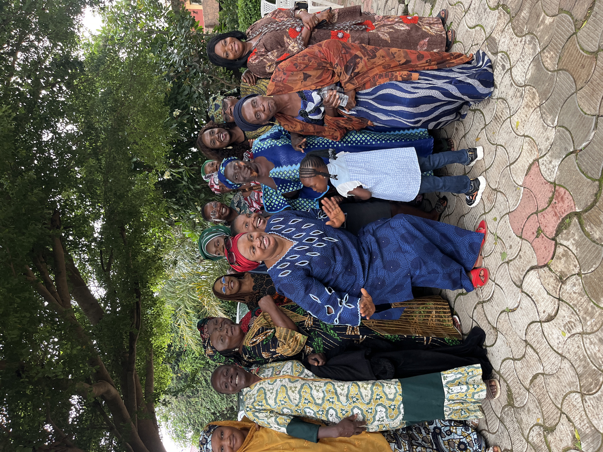 In a medium shot, a group of approximately fifteen women and one young girl are gathered outdoors, posing for a photograph under the dappled shade of a large tree. The women are dressed in vibrant, traditional African attire, featuring bold patterns and colorful fabrics. Many of them are wearing headscarves. The group appears to be in good spirits, with several individuals smiling and one woman at the forefront in a blue outfit is actively gesturing with her hands. The young girl, wearing a light blue and white checkered dress and dark leggings, stands in the front center of the group, looking towards the camera. The ground beneath them is paved with interlocking concrete tiles, some of which show patches of green algae. Behind the group, lush green foliage, including palm fronds, and a hint of a red-brick building are visible. The lighting suggests it is daytime, with sunlight filtering through the tree canopy.