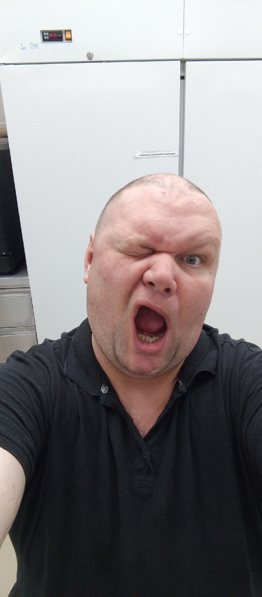A man with a shaved head and blue eyes winks and yells, his mouth wide open, revealing his teeth. He wears a black collared shirt. The background is a plain white wall. The scene appears to be indoors, possibly in a laboratory or industrial setting, given the presence of a white refrigerator-like unit with a digital display and buttons in the upper left corner of the frame. A partial view of a black object on the far left suggests it might be a piece of equipment or furniture. The image is a close-up selfie, capturing the man's expressive face and upper torso. No specific location in Bugry, Russia is discernible from the visual information provided.