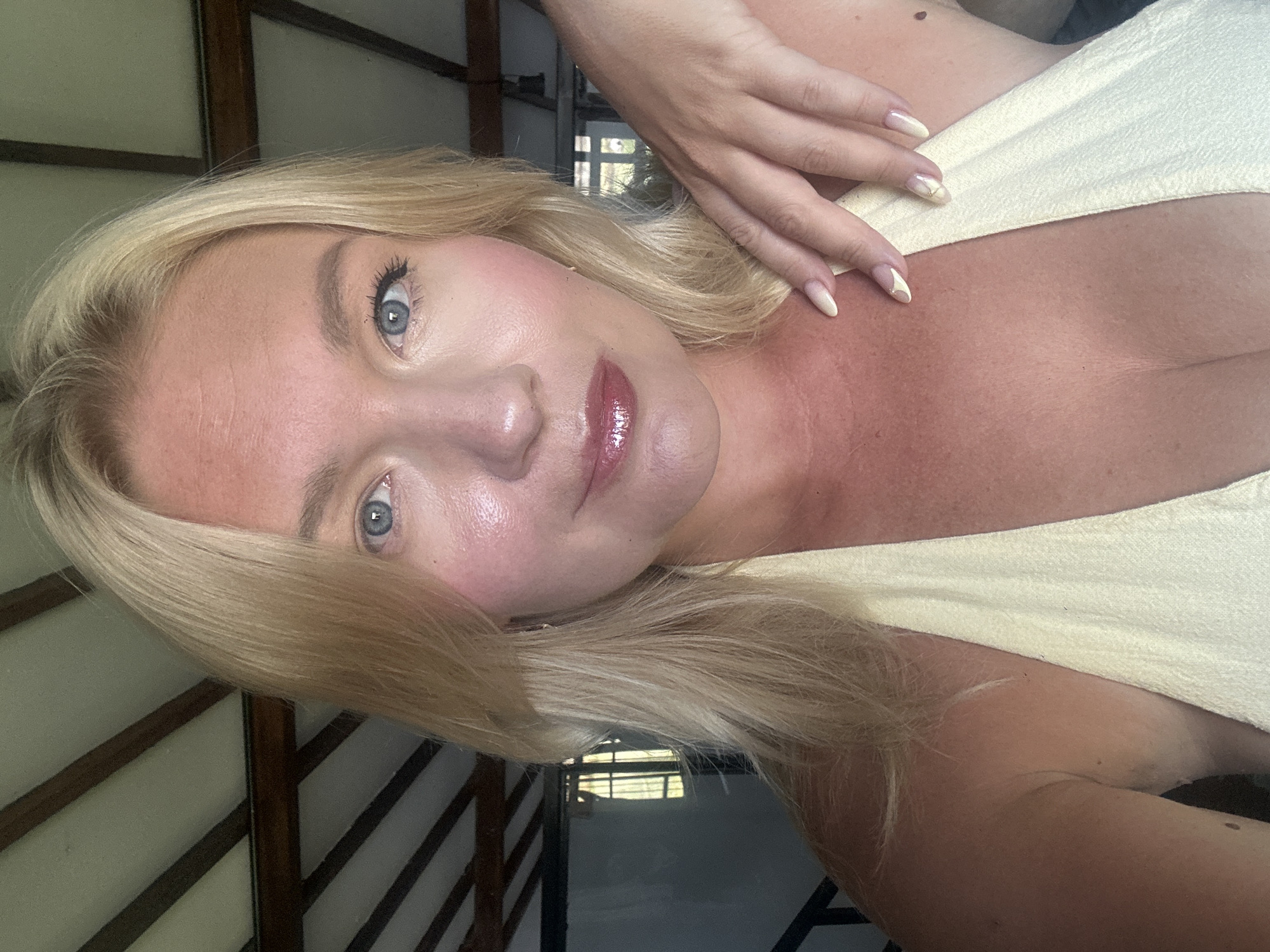 A close-up, slightly high-angle selfie features a blonde woman with blue eyes and fair skin, wearing a light yellow halter top. Her skin appears slightly flushed and possibly sun-kissed. She has on lip gloss and subtle eye makeup. Her left hand is resting on her right shoulder, with painted nails. In the background, a wooden-beamed ceiling with white panels is visible, suggesting an indoor setting with natural light. The overall impression is casual and relaxed. The image was taken in North Kuta, Indonesia.