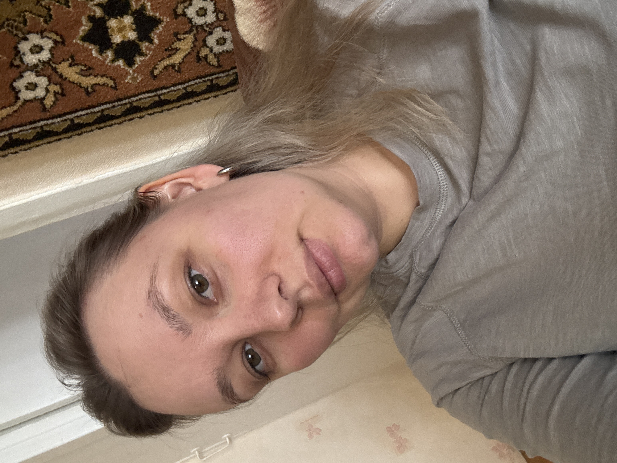 The image captures a close-up, chest-up portrait of an individual, likely female, indoors in Chaikovsky, Russia. The subject, appearing to be in her late 20s to early 30s, is fair-skinned with light brown to blonde hair, parted on the left and falling loosely. Her medium brown eyes are open and directed at the camera. Her eyebrows are light brown and naturally shaped. She exhibits a subtle, relaxed facial expression with closed lips. The left ear is visible, adorned with a small, silver-colored hoop earring. She is wearing a light grey, long-sleeved shirt with distinct stitching visible on the shoulder and collar. The background on the left features a light-colored wall with faint pink floral patterns and a white, angular fixture at the upper left. On the right, a dark brown textile or rug with intricate floral and geometric patterns in off-white, black, and gold hangs vertically. A narrow, light-colored vertical strip separates the wall and the patterned textile. The scene is illuminated by ambient interior light.