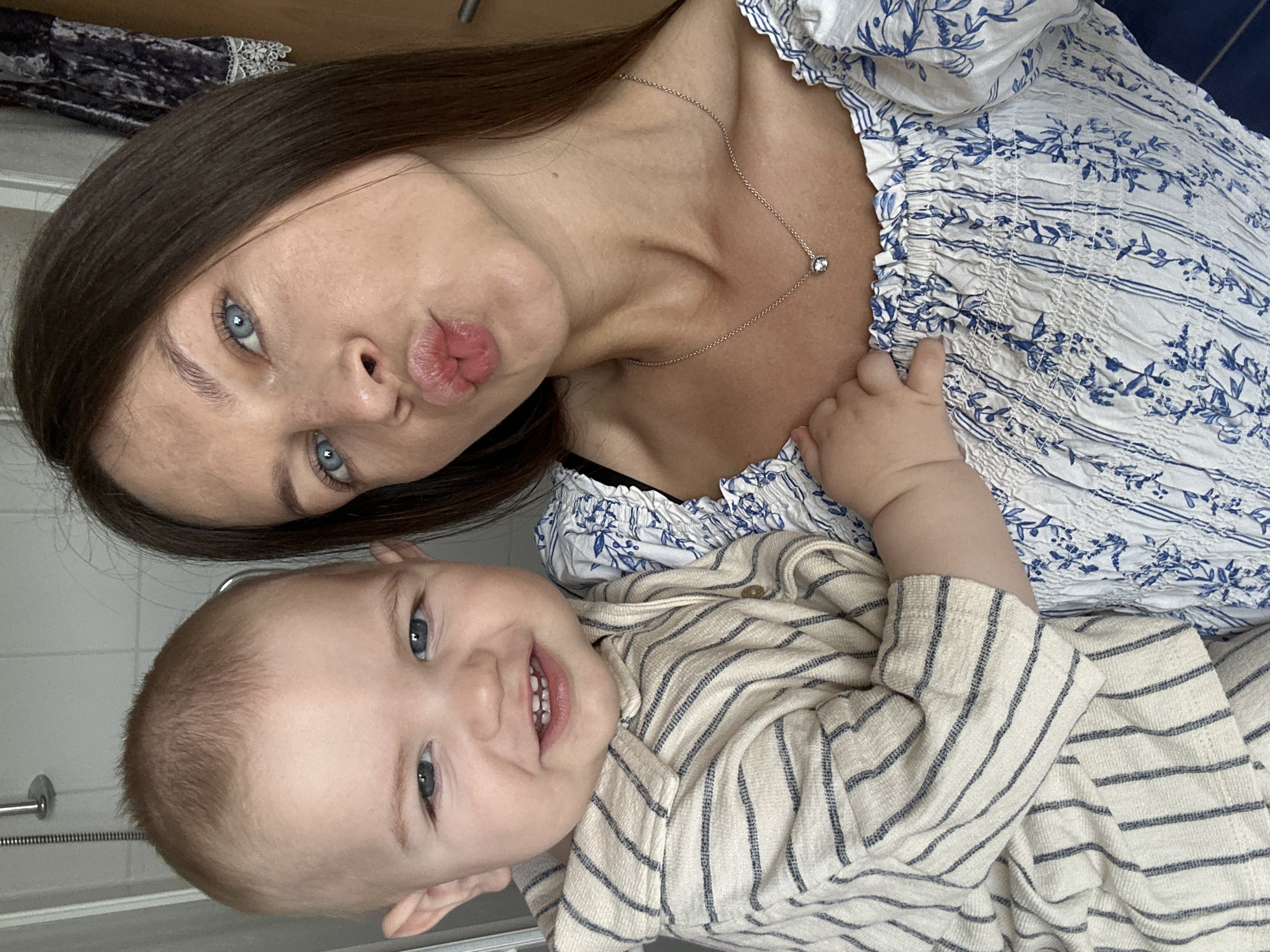 A selfie shows a woman and a baby. The woman has long brown hair, blue eyes, and is making a kissing face. She wears a white blouse with blue floral patterns and a silver necklace. The baby has blond hair, blue eyes, and is smiling, wearing a striped beige shirt. They appear to be indoors, possibly in a bathroom, indicated by the shower fixture and tiles in the background. The photo does not provide enough information to determine if this was taken in Niederquembach, Germany.