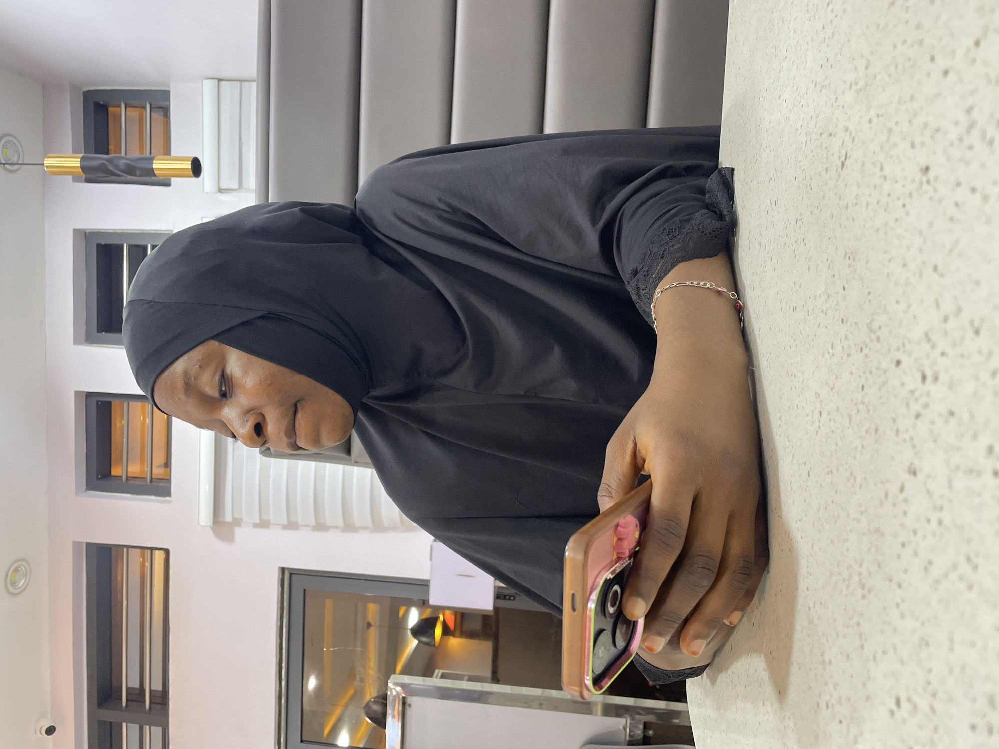 A woman wearing a black hijab is seated at a white countertop, holding a cell phone with a pink and gold case. She appears to be looking at the phone. The location is likely in an indoor setting in Jos, Nigeria, as evidenced by the style of windows and the local cuisine. Behind her is a gray, padded wall panel. The background also reveals interior lighting fixtures.