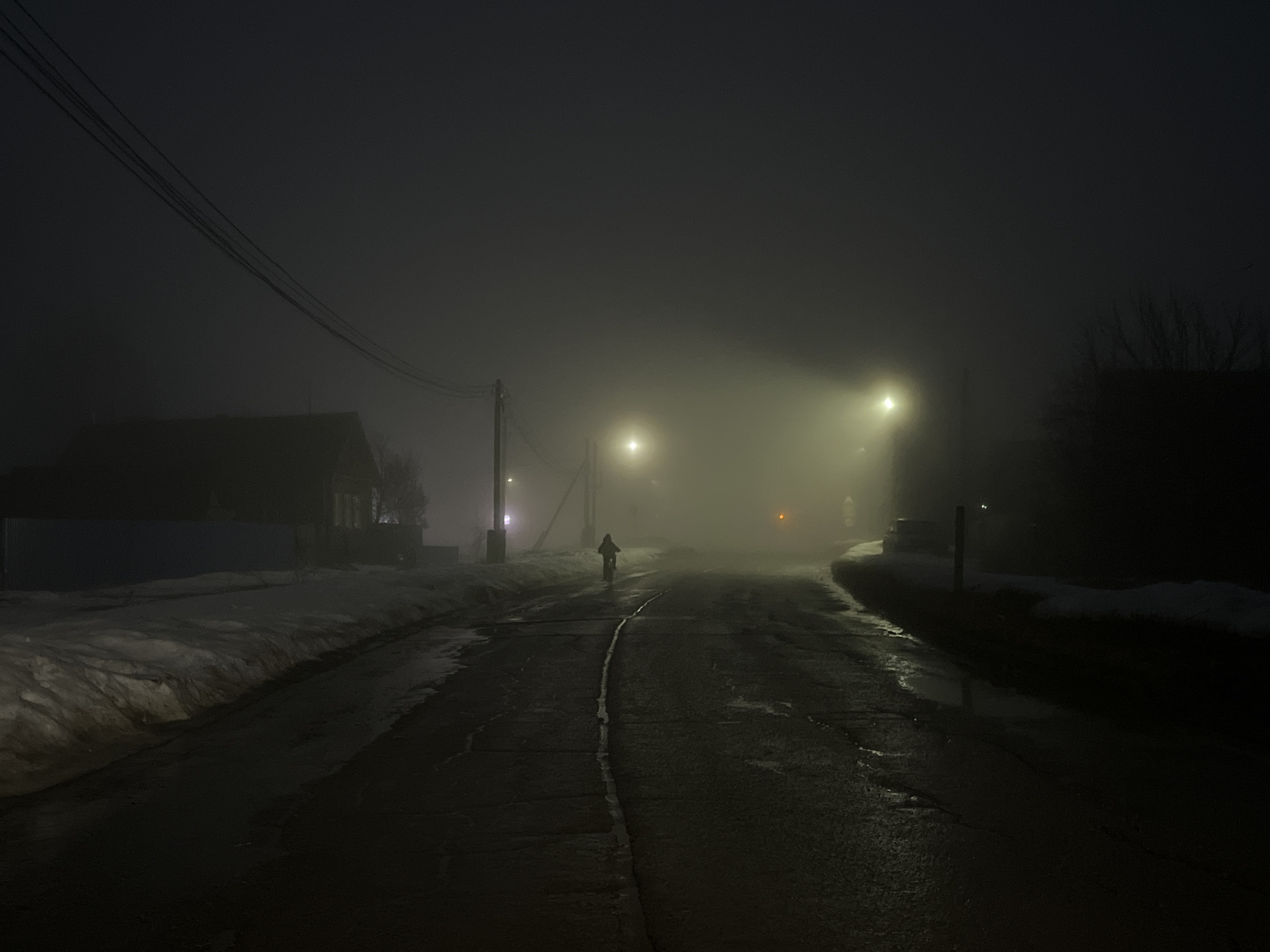 The photograph captures a dimly lit street scene at night in Сосново, Russia. The atmosphere is thick with fog, obscuring much of the surroundings. A lone figure on a bicycle is seen riding away from the viewer, their silhouette illuminated by distant streetlights. On either side of the road, patches of snow remain. The road surface is dark and appears wet, reflecting the faint light. Buildings and trees are partially visible on the sides, but their details are lost in the fog. Overhead, power lines stretch across the sky. The overall impression is one of a quiet, desolate, and foggy night.