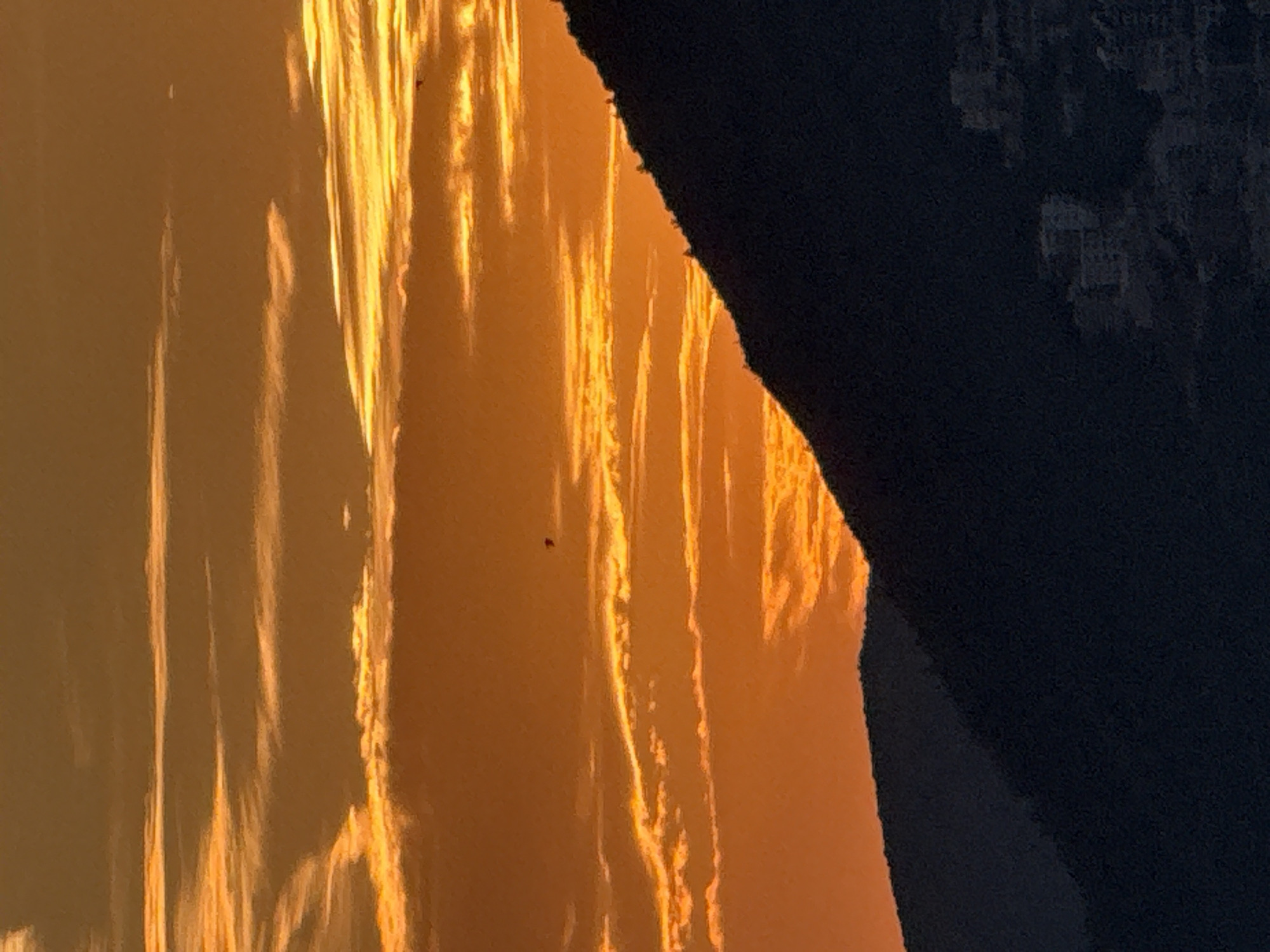 An orange sunset casts a warm glow over the sky, illuminating wispy clouds. In the foreground, the dark silhouette of a mountain range stretches across the frame. The lower slopes of the mountain are dotted with the faint lights of a town, suggesting the presence of buildings and infrastructure. A small, dark object, possibly a bird or kite, is visible in flight against the vibrant sky. The scene appears to be set in the city of Pržno, Montenegro, offering a serene natural landscape juxtaposed with hints of human habitation.