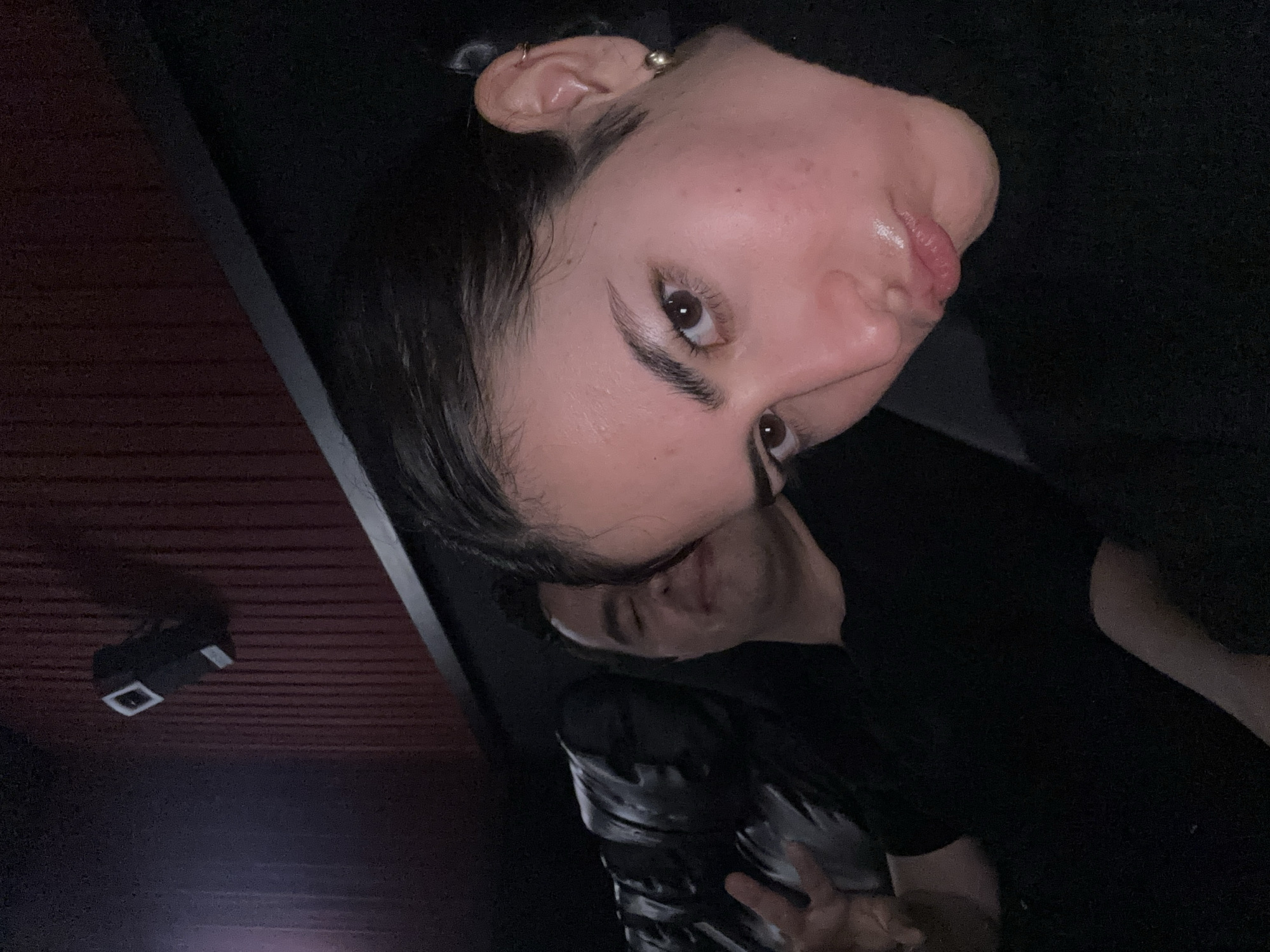 A low-angle close-up shot of a fair-skinned woman with dark hair pulled back, showing her face, particularly her right eye and right ear with a small gold hoop earring. She's wearing dark clothing. Behind her and partially obscured is another person in darker clothing. The ceiling is visible in the upper left of the frame, showing wood paneling and a small rectangular light fixture. The location cannot be determined from this image.