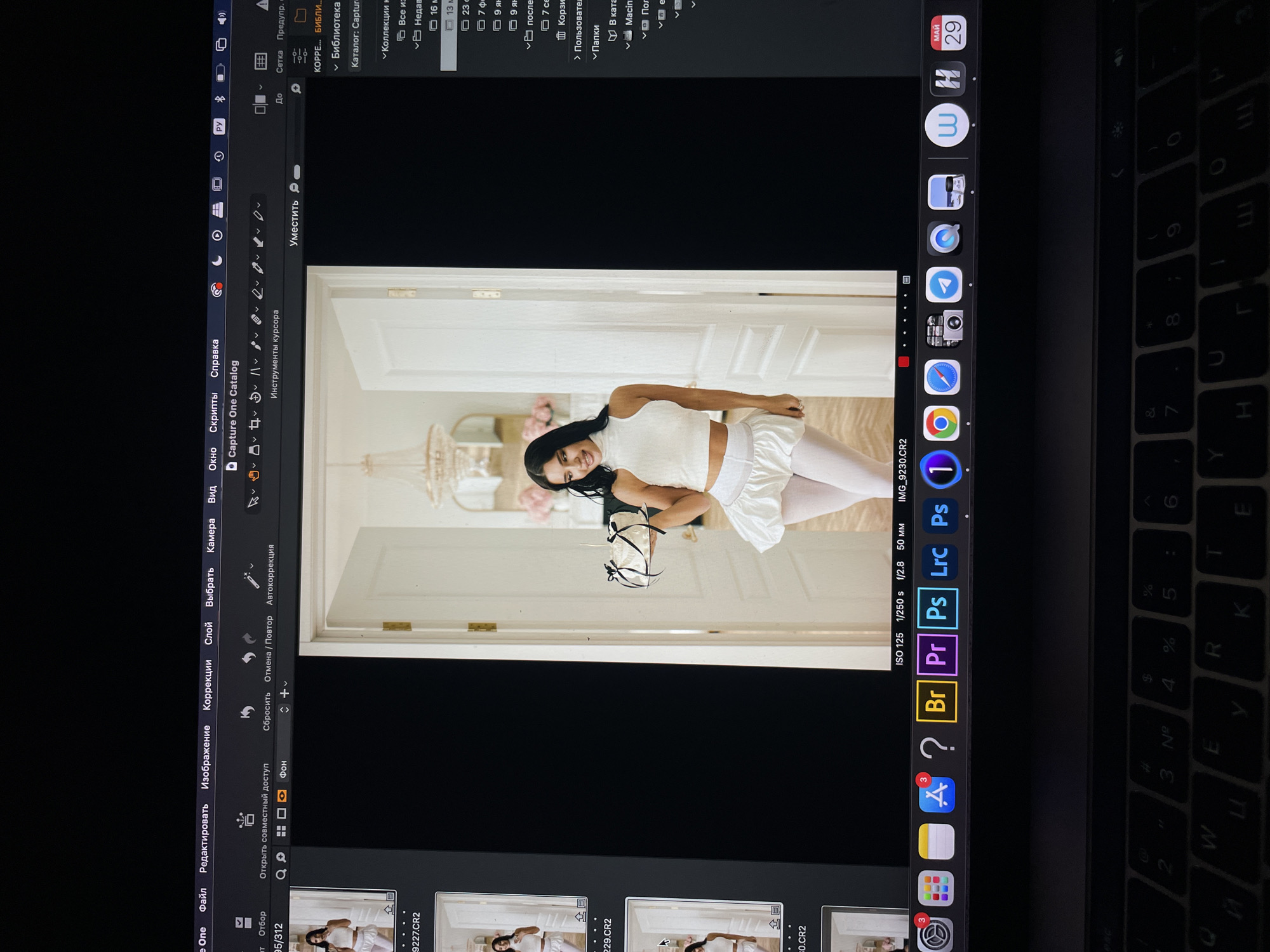The image is a photograph of a laptop screen displaying a photo editing interface. The photo editing software is Capture One, with a portrait-oriented photograph in the main display area. The photograph features a young woman in a white outfit, standing in a doorway. The doorway appears to lead into a room with soft, pastel decor. The operating system appears to be macOS, with the dock visible at the bottom of the screen. App icons such as Safari, Photos, App Store, and Adobe products (Bridge, Premiere Pro, Photoshop, Lightroom) are visible. The date "МАЙ 29" is displayed in the top right corner of the screen, which translates to "May 29." A portion of the laptop's keyboard is also visible, with Russian letters on the keys. The location within the city of Kazan, Russia, is not discernible from the content of the image.
