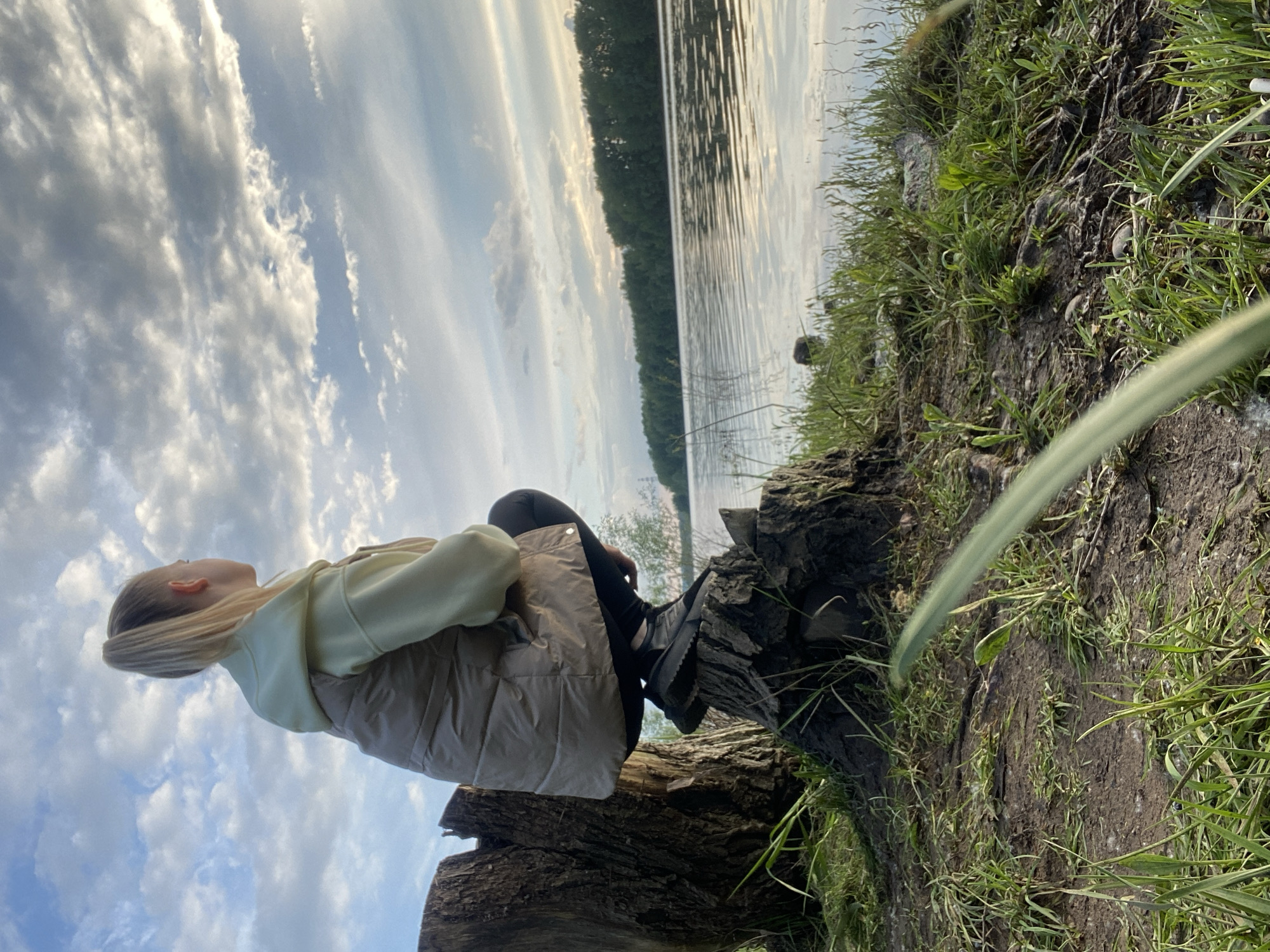A young woman is sitting on a tree stump near a lake in what appears to be a natural environment. The lake and surrounding forest suggest a location outside of an urban center. While the image is rotated, it's possible to discern the sky is filled with clouds, creating a dynamic backdrop. The woman is wearing a hooded sweatshirt and a vest, and appears to be wearing boots. The ground around the stump is covered in grass. The location in the city of Chaikovsky, Russia cannot be confirmed.