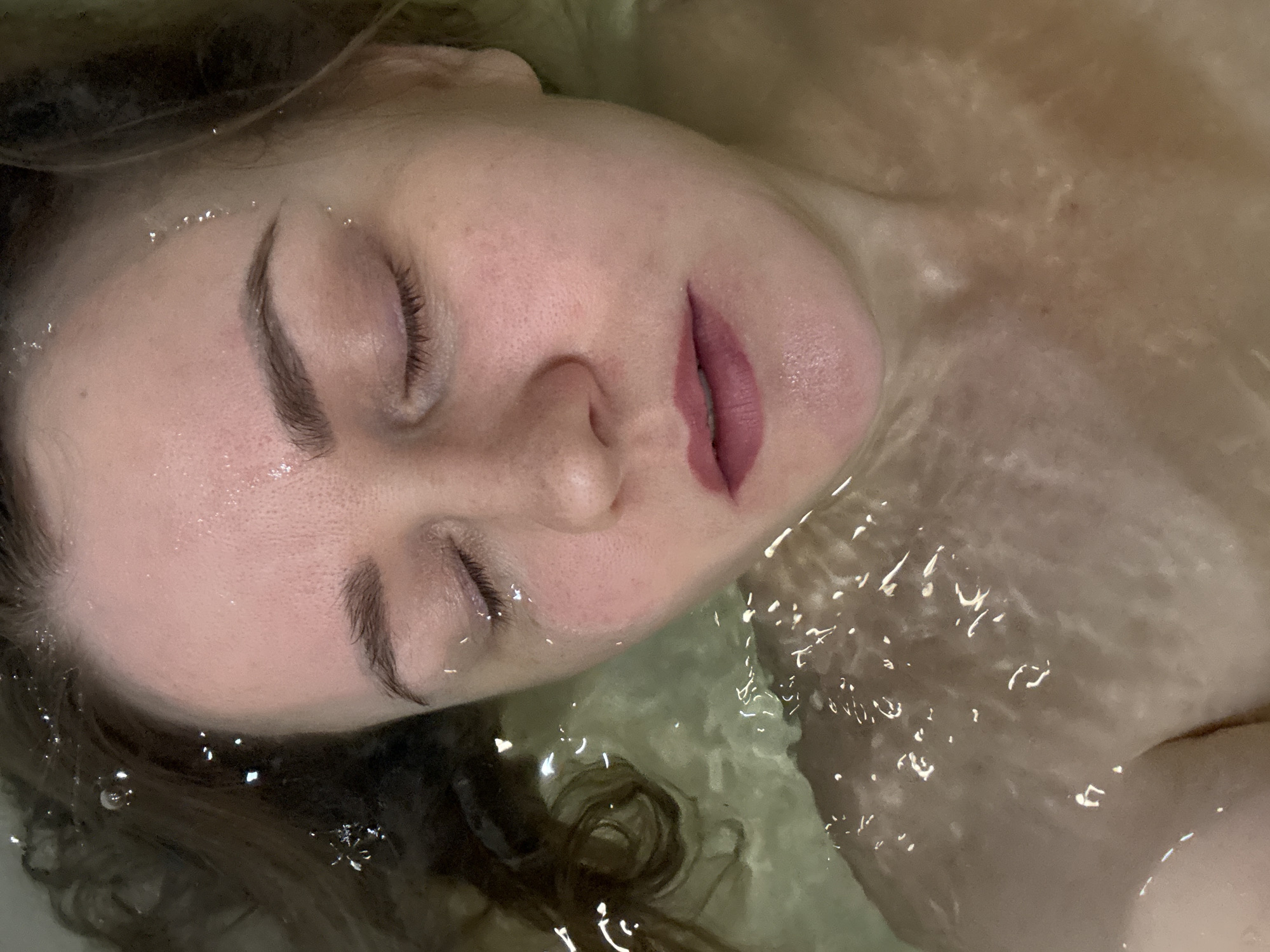The image captures a close-up, high-angle view of a woman floating in shallow, translucent water, likely in a bathtub or a pool, in Chaikovsky, Russia. Her head and upper chest are visible, with her face turned slightly to the right, eyes peacefully closed.
**Subjects**: The primary subject is a young woman. Her fair skin shows a few subtle blemishes or pores, and her eyebrows are dark and well-defined. Her lips are a soft reddish-pink shade. Her dark hair is partially submerged and fanned out around her head.
**Activity or event taking place**: The woman appears to be resting or relaxing, fully immersed in the water up to her shoulders, with only her face and parts of her hair breaking the surface. Her closed eyes convey a sense of calm and serenity, or perhaps contemplation.
**Notable details**:
* **Emotions**: The closed eyes and relaxed facial features suggest a state of peace, tranquility, or deep relaxation.
* **Water**: The water is clear, with subtle greenish-brownish hues, and shows numerous ripples and light reflections, creating sparkling highlights across her forehead, eyelashes, and hair. Small water droplets are visible on her skin and lashes.
* **Lighting**: The lighting is soft and diffused, highlighting the contours of her face and the glistening water.
* **Details from crops**: The close-up crops emphasize the texture of her skin, the individual hairs of her eyebrows and eyelashes, and the detailed reflections of light on the water's surface, particularly around her eyes and hair. No specific time of day or weather is indicated, but the indoor setting is implied.
**Visible text**: There is no visible text in the image.