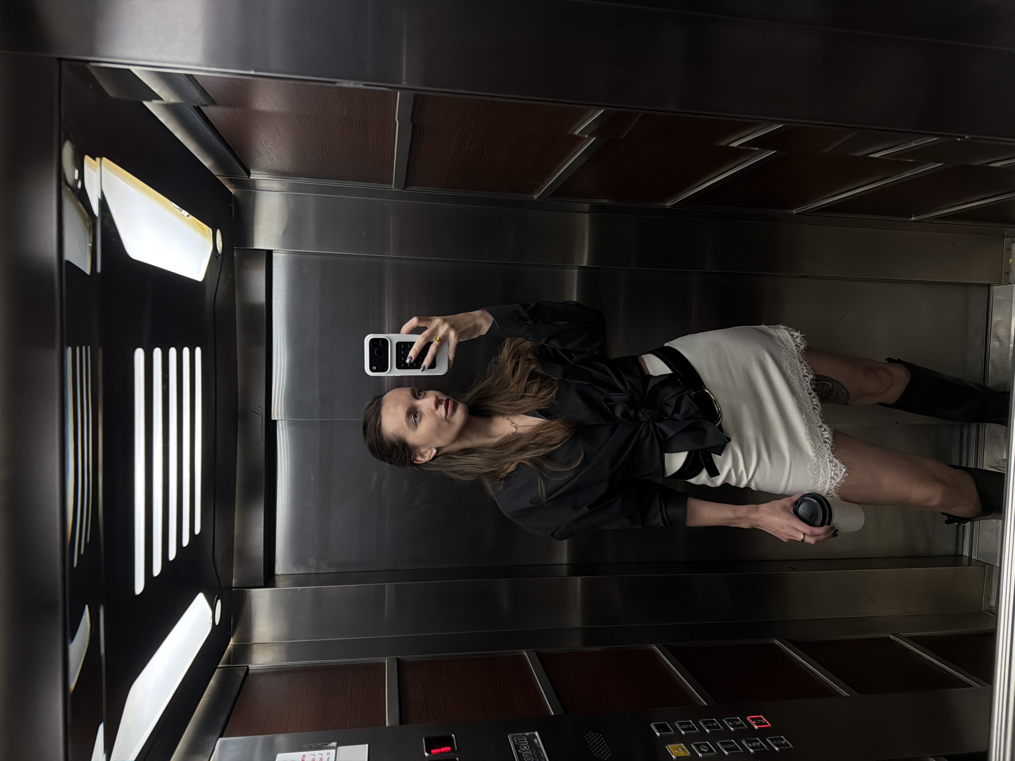 The image shows a woman inside an elevator. She is holding a smartphone in her right hand, seemingly taking a selfie in the mirror on the back wall of the elevator. She also holds a disposable coffee cup in her left hand. The woman is wearing a black top, a white skirt with lace trim, and black boots. The elevator's interior is a mix of stainless steel and brown wood paneling. A control panel with buttons is located at the bottom of the elevator, and an overhead light is visible at the top. The location in the city of Budva, Montenegro cannot be determined from the image.