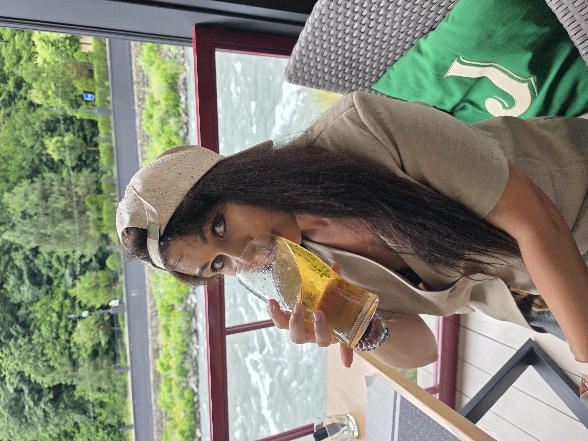 A woman is seated at a table with a glass of beer in her hand, drinking it. She is wearing a beige blazer and a beige cap with rhinestones. She is also wearing bracelets on her left wrist. In the background, a river and trees are visible. The scene is outdoors on a balcony or terrace.