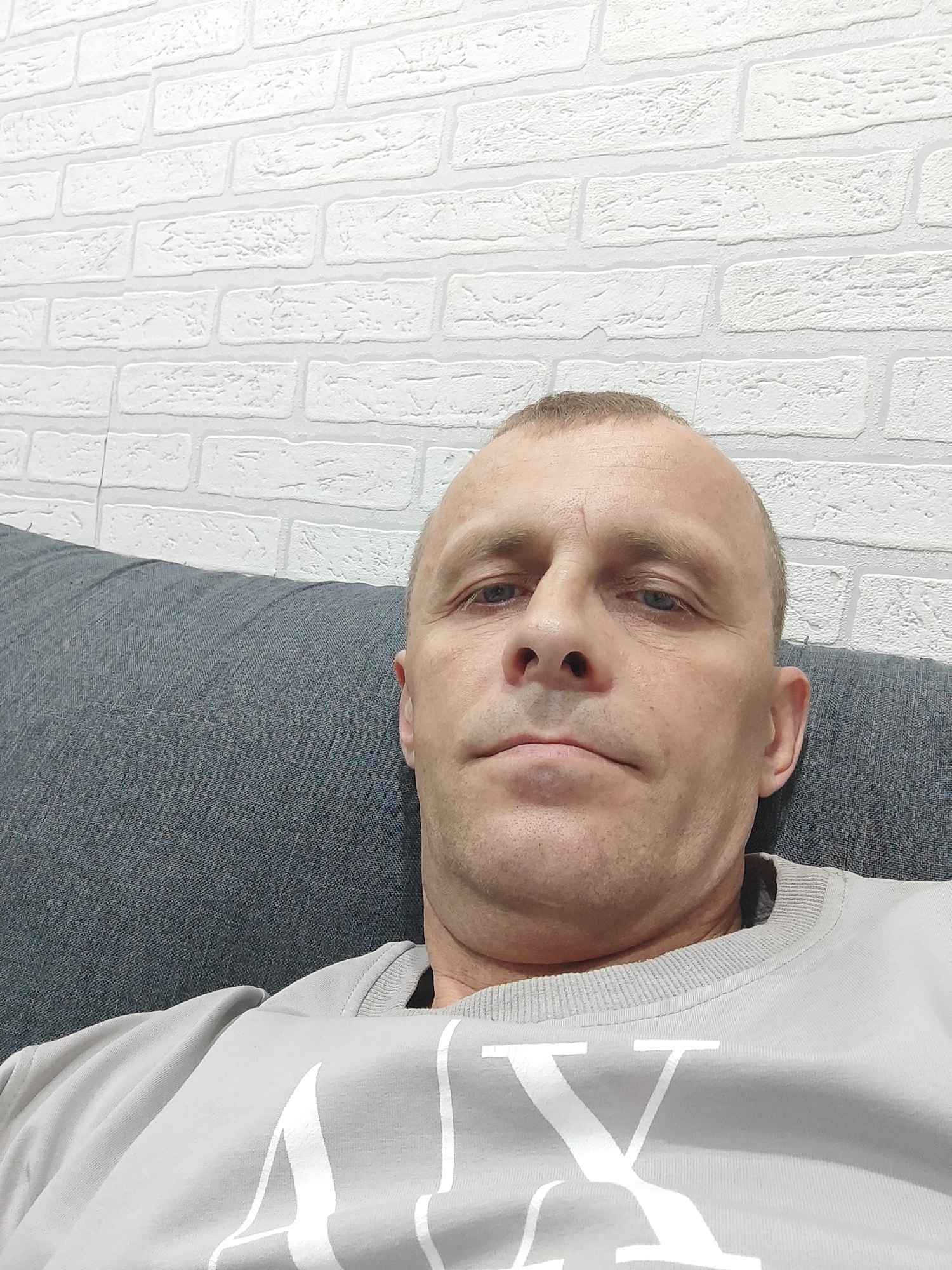 A middle-aged Caucasian male with fair skin, short brown hair, and blue eyes is depicted in a close-up, low-angle shot. He has a neutral expression. He is resting against a gray upholstered surface, likely a sofa or headboard. The man is wearing a light gray sweatshirt with a large white logo (letters "AX"). The background is a white brick wall. There is no indication of the location being Malaryta, Belarus. The image features no evident activities or interactions.
