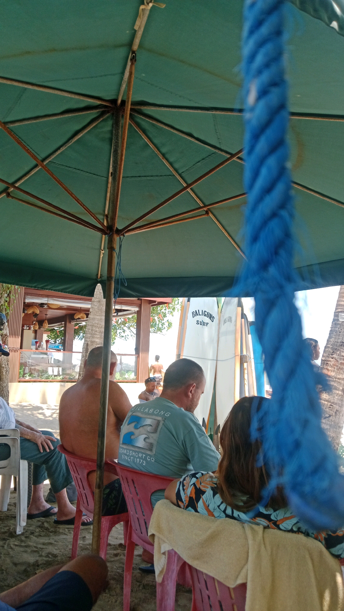 An outdoor scene at a beachside establishment in Kuta, Indonesia, shows several people seated beneath a large, green umbrella. The ground is sandy. In the foreground, a thick, blue rope hangs loosely, obscuring part of the view. Two men are seated on red plastic chairs, facing away from the camera. The man on the left is bare-chested. The man on the right wears a light blue t-shirt with a wave design and the text "BILLABONG BOARDSHORT CO. SINCE 1973". Another person, whose back is to the camera, sits to the right of the man in the t-shirt, wearing a patterned top. Partially visible to the left, a person wearing blue pants and dark sandals sits on a white chair. In the background, behind the seated individuals, are several surfboards leaning against a structure with an open-air section. People can be seen walking in the distance.