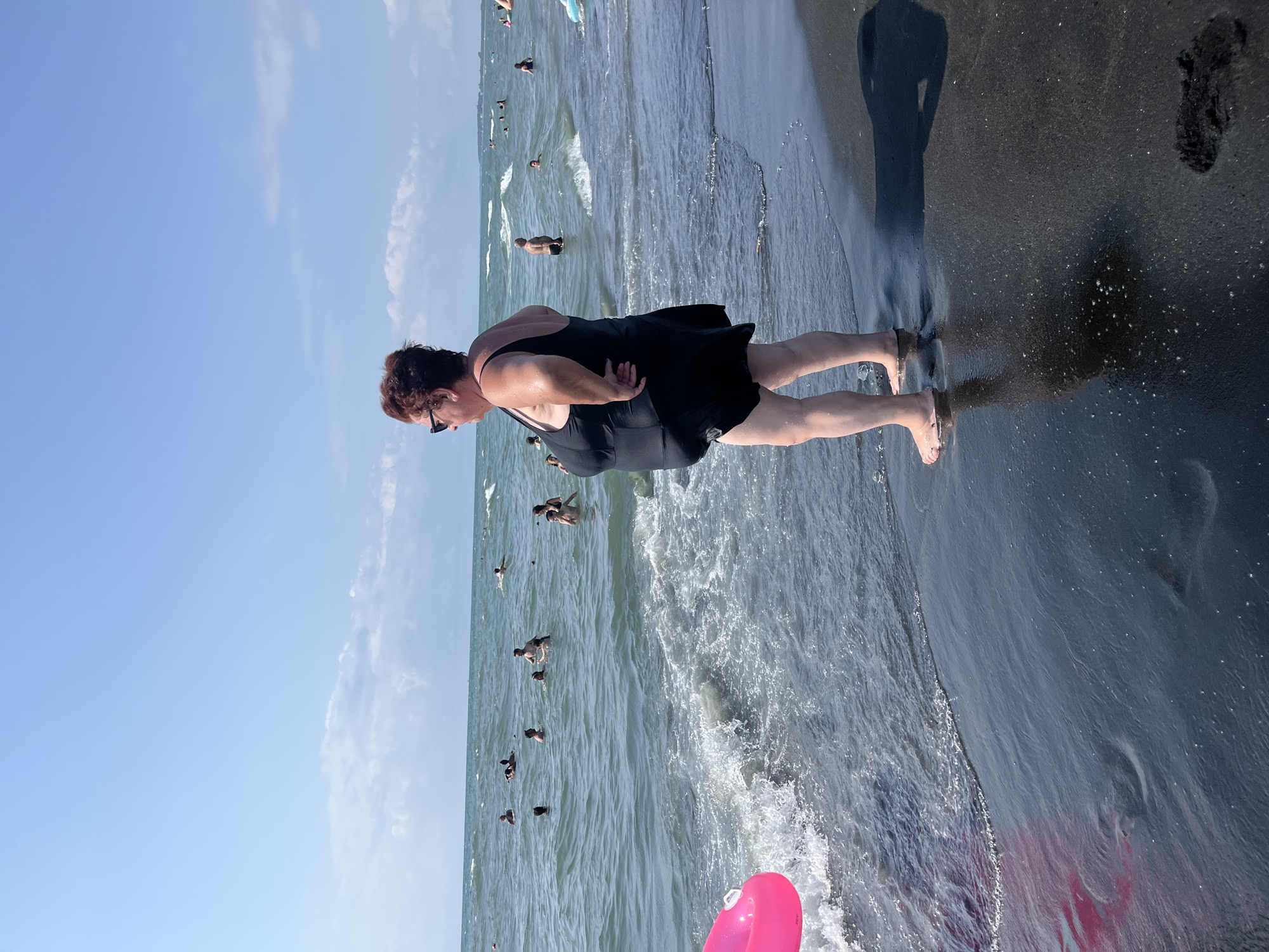 A woman wearing a black one-piece swimsuit and sunglasses stands on a black sand beach, facing right with her hands on her hips. She is looking out at the sea, where other people are swimming and wading in the water. A pink inflatable ring is partially visible in the foreground on the left. The sky is blue with scattered white clouds. The scene appears to be in Ureki, Georgia, known for its magnetic black sand beaches.