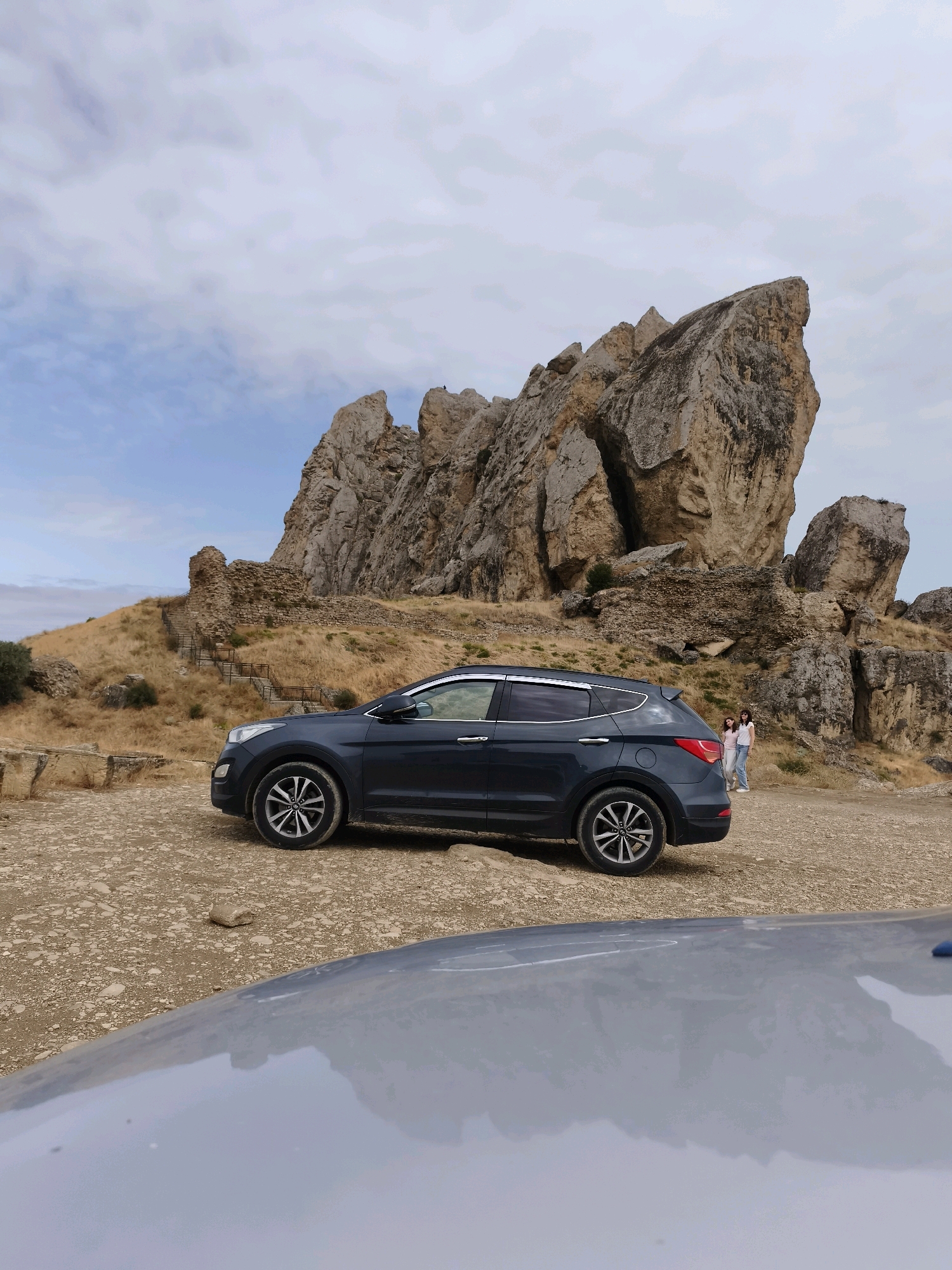 A dark blue SUV is parked on a dirt road in front of a rocky, mountainous landscape. Two individuals are visible standing in the distance near the rocks. The terrain is dry and covered with sparse vegetation. The sky is overcast with clouds. In the foreground, the reflection of another vehicle is visible on a shiny surface. The scene appears to be an outdoor location, possibly a historical or natural site.