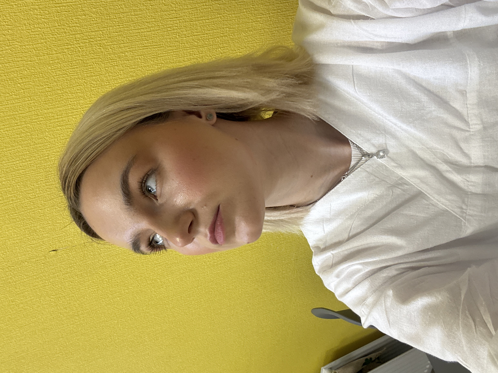 A woman with short blonde hair is indoors, standing or sitting with a yellow wall behind her. She is wearing a white shirt and a delicate silver necklace with a small, clear pendant. The lighting in the room is bright. The photo is taken at an angle that slightly tilts the subject. The location in Saint Petersburg, Russia is not identifiable in this image.