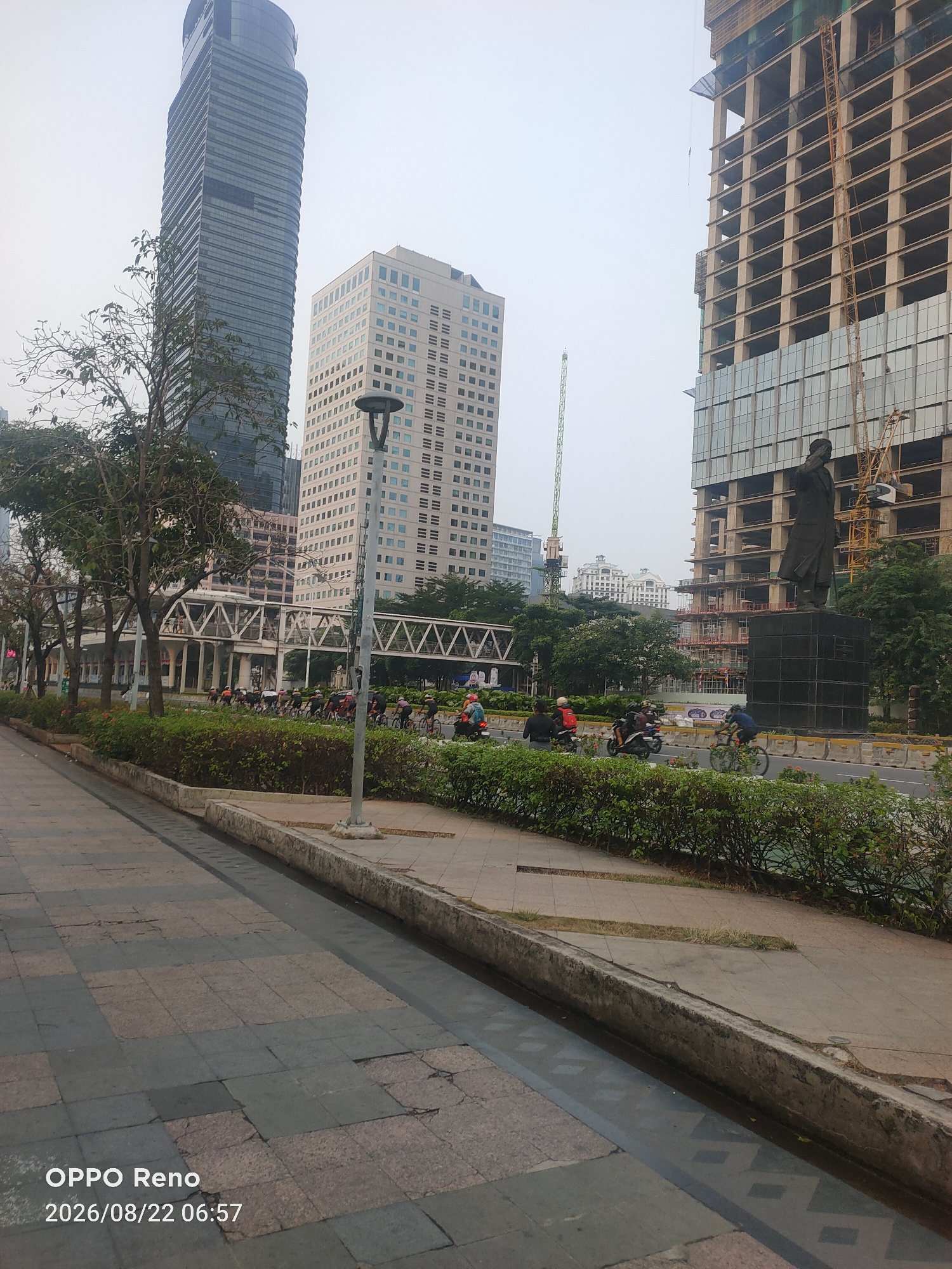 A street scene in an urban area features a sidewalk on the left, a road with vehicles and cyclists, and several buildings in the background. The sidewalk is paved with gray and pinkish square tiles. Adjacent to the sidewalk is a concrete channel, possibly a planter or drainage system, filled with water reflecting diamond shapes. Beyond this channel is a row of green bushes.
The road has multiple lanes and shows traffic. Several individuals are riding bicycles, and some are on motorcycles. A group of cyclists is visible on the road further down.
In the background, there are multiple tall buildings. On the far left is a sleek, dark glass skyscraper. To its right is a lighter-colored rectangular building with many windows. In the center background, a construction crane is visible next to a tall building under construction. To the right of that, a large, dark statue stands on a pedestal in front of a building with exposed concrete beams and glass panels, also appearing to be under construction, with cranes nearby.
Overhead, the sky is overcast and pale. The overall scene suggests a bustling urban environment with ongoing development. The timestamp at the bottom left corner of the image reads "OPPO Reno 2026/08/22 06:57".