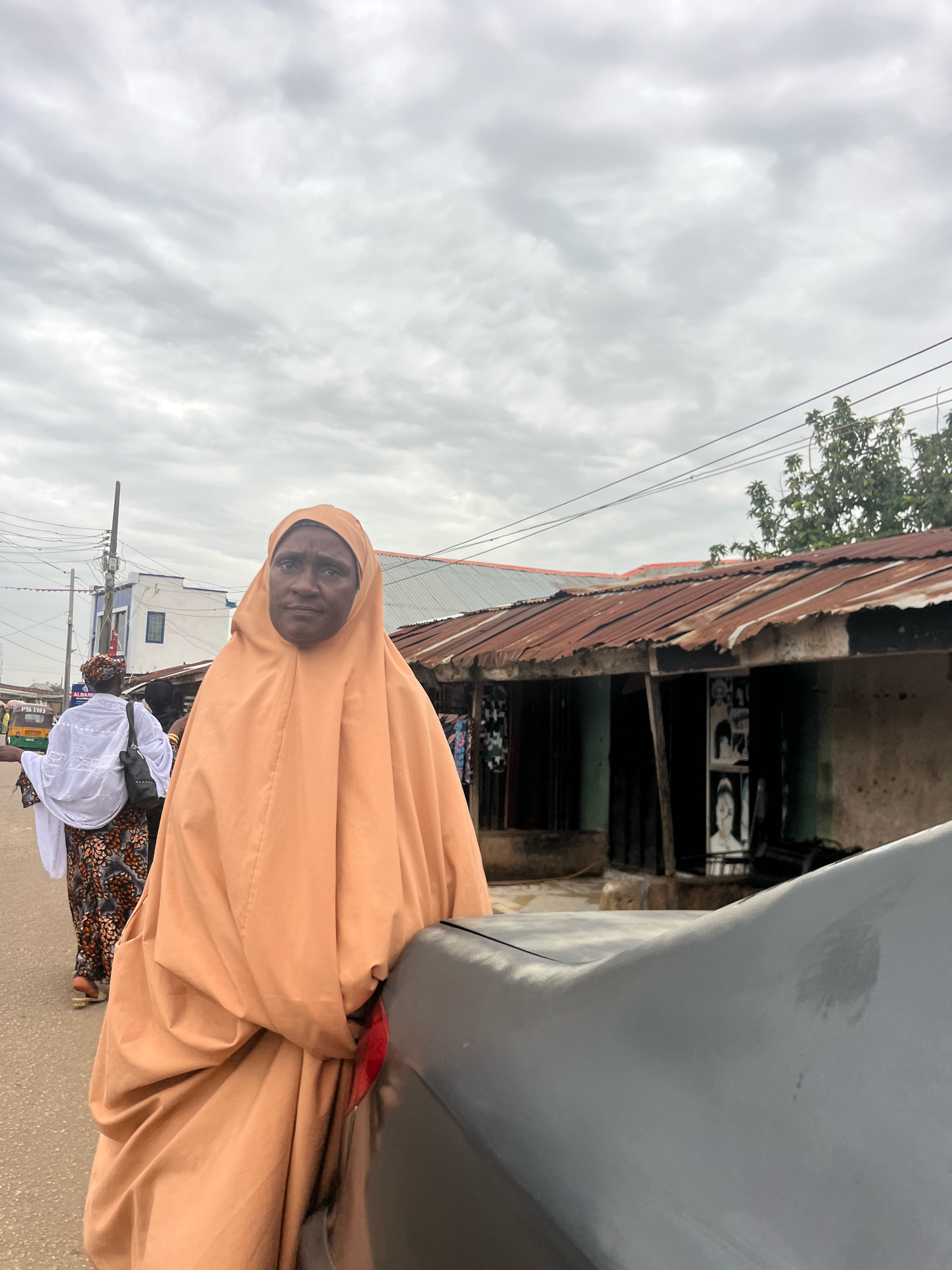 A woman wearing an orange hijab and traditional attire stands in a street in Jos, Nigeria. She is in the foreground, her body angled slightly away from the viewer. Behind her, other people are visible walking down the street, some in traditional clothing as well. In the background, there are buildings with corrugated iron roofs, typical of market areas or residential areas in Nigerian cities. A small vehicle and utility poles are also present, indicating an urban environment. The sky is overcast, suggesting a cloudy day. The scene captures a moment of everyday life in a bustling Nigerian city.
