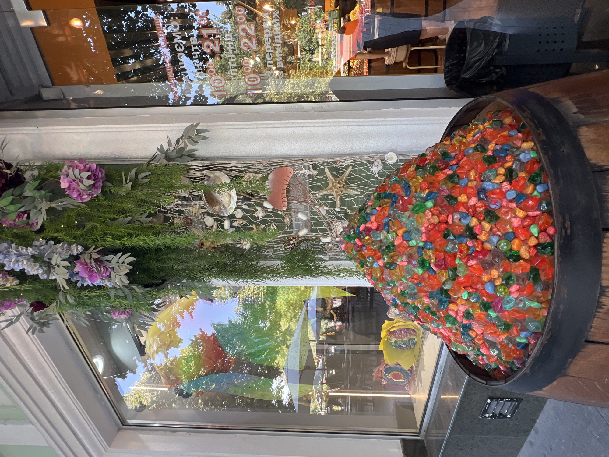 The image depicts the storefront of a business in Odesa, Ukraine. In the foreground, a large wooden barrel is filled with a mound of colorful, translucent decorative stones. The barrel has a dark metal rim. To the right of the barrel, a black trash can with a mesh design is visible, and further right, a person's legs are seen wearing white shoes and a dark skirt or shorts, standing near a stool with a bucket on it.
The storefront features a large display window filled with an assortment of items. In the window, a display of colorful decorations, including what appears to be a large yellow fan-like object and other smaller colorful items, is visible. Beyond the display, the window reflects an outdoor scene with trees, umbrellas, and possibly tables and chairs of an outdoor seating area.
Hanging above the barrel and partially obscuring the display window is a decorative arrangement of artificial greenery, pink and purple flowers, and various oceanic-themed decorations including shells, a jellyfish, and a starfish, all suspended from a net-like material.
To the right of the display window is a glass door, also reflective, showing more of the outdoor environment and some text on the glass, which appears to be operating hours. Part of the text visible reads "10:00 - 21:00" and "10:00 - 22:00", suggesting business operating hours. The overall impression is a vibrant and somewhat eclectic street-level commercial facade.