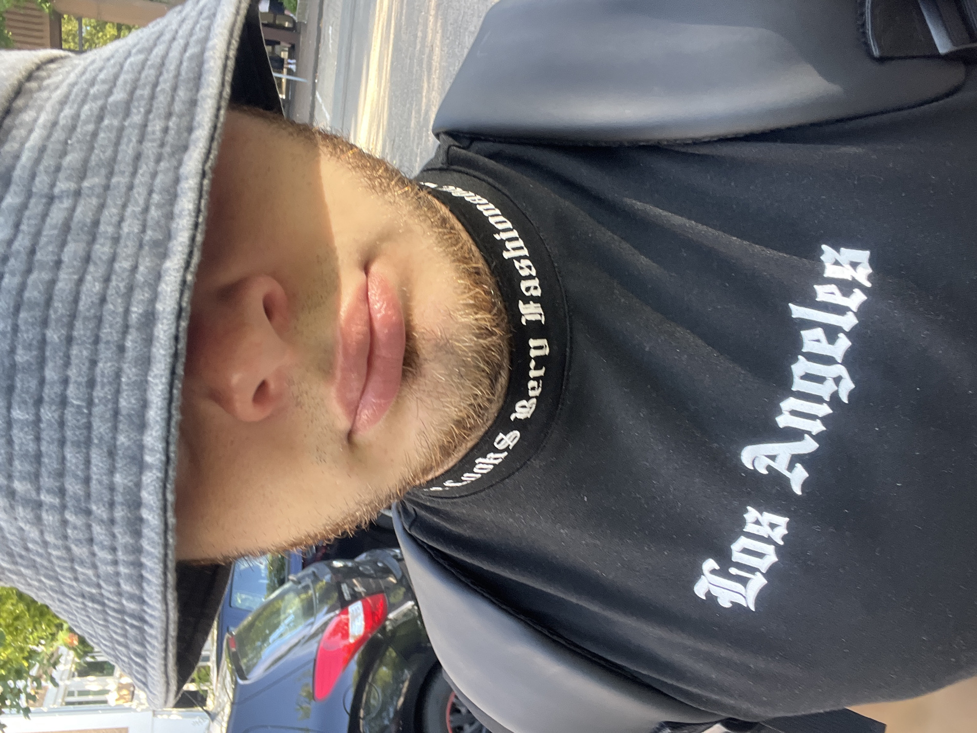 A close-up, waist-up shot shows a person wearing a gray bucket hat and a black t-shirt with white lettering. The hat covers the upper half of the face, obscuring the eyes and nose, but revealing the lips and chin, which are covered in light stubble. The t-shirt has two lines of text; the top line, encircling the neck, reads "Looks Very Fashion" in a gothic font. The bottom line, across the chest, reads "Los Angeles" in a similar font. The person is wearing a black backpack over their shoulders. The background shows a parked car and a street with a sidewalk. The lighting suggests it is daytime, with sunlight casting shadows.