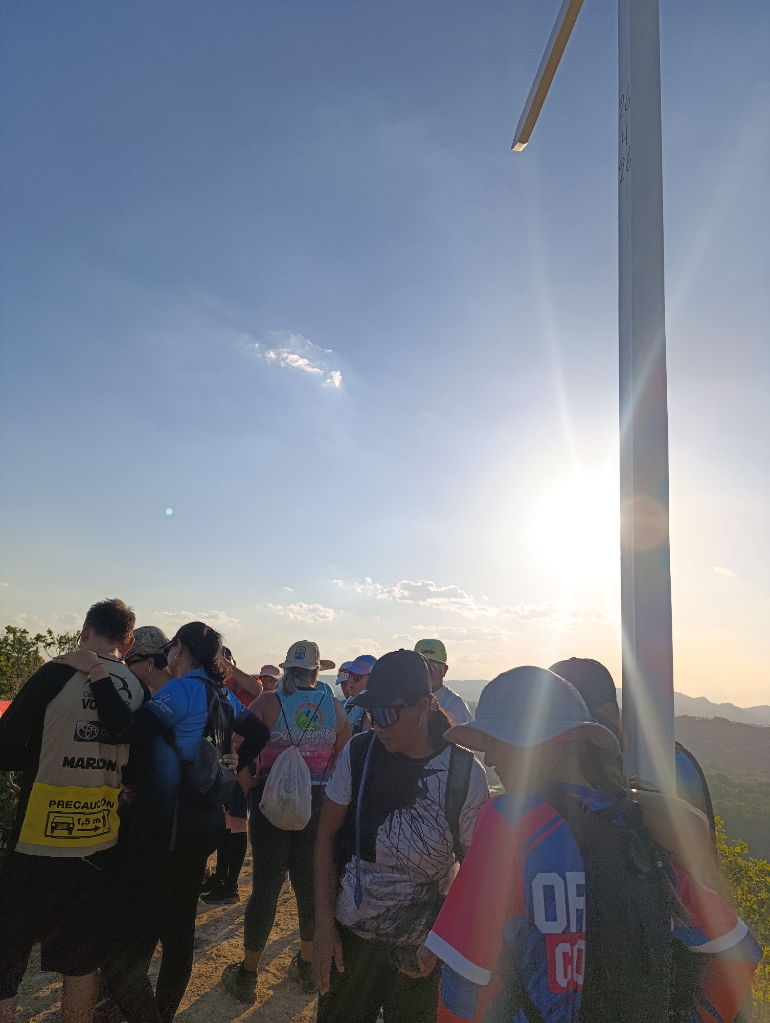 A daytime scene shows a group of people congregated near a tall, white cross at what appears to be a high-altitude location in Tinaquillo, Venezuela. The people are wearing casual clothing, including t-shirts, hats, and backpacks. The sun is bright, casting a glare in the vicinity of the cross. The cross has numbers "26 14 26" written on it. The background reveals a mountainous landscape. A person on the left has a vest with the word "PRECAUCION" written on it, along with an image of a car.