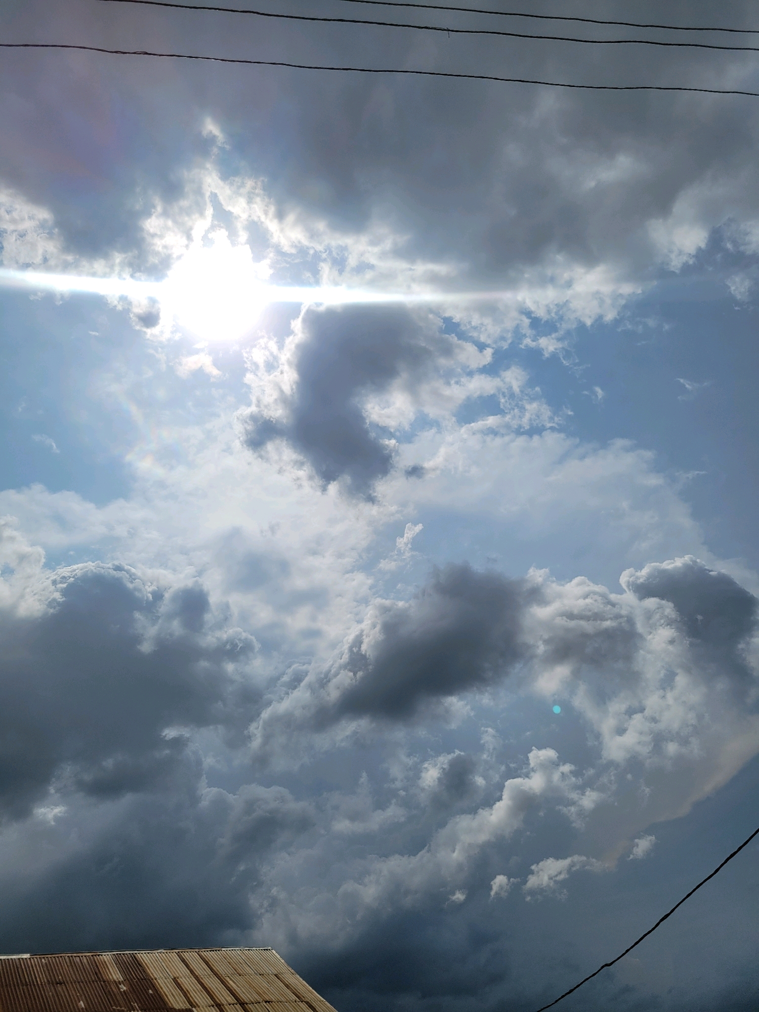 The image captures a dramatic sky above Jos, Nigeria, dominated by a mix of dark, ominous storm clouds and bright, sunlit patches. The sun is a powerful, glaring presence, creating a lens flare that streaks horizontally across the upper portion of the frame, illuminating the edges of some clouds with a brilliant white light. Below, the clouds vary in density and color, ranging from deep grey to lighter white, suggesting turbulent weather.
In the foreground, the corrugated metal roof of a building is visible in the lower left corner, showing signs of rust and age, indicating an outdoor scene. The roof is angled upwards, suggesting it belongs to a structure situated on a slight incline or viewed from a low vantage point. Thin, dark lines, likely overhead wires, are stretched across the top of the image, further grounding the celestial view in a human-made environment. A faint, almost iridescent rainbow-like effect is visible in the upper left quadrant, a subtle detail amidst the dramatic cloud formations. The overall impression is one of impending weather change, with the harsh sunlight battling the encroaching storm clouds.