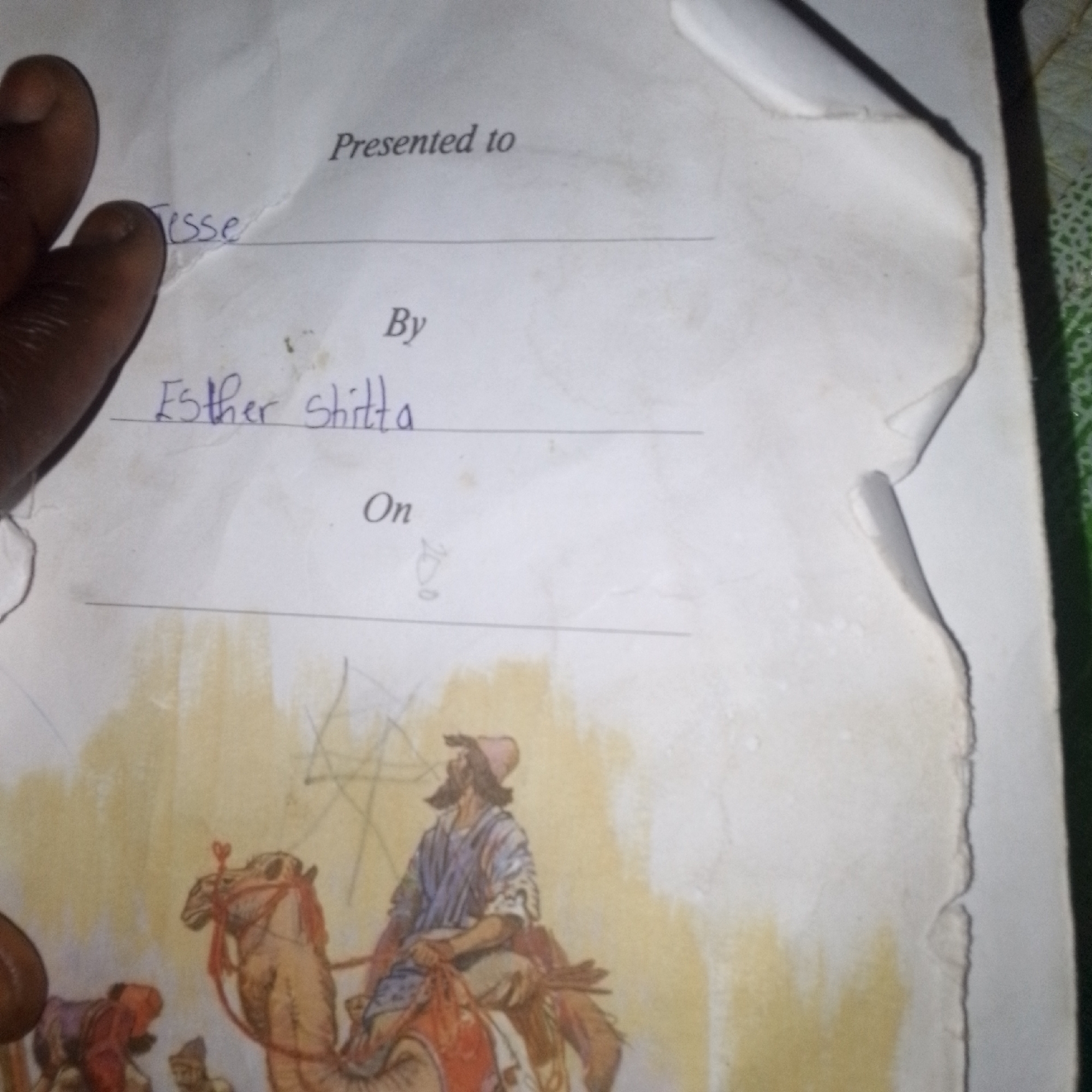 The image shows a piece of paper with handwriting on it, indicating a certificate or award. The text reads "Presented to" followed by a line for a name, then "By" followed by "Esther shitta", and finally "On" with a line for a date. Below this text, there is a colored illustration of a man riding a camel, with other figures and camels in the background. The paper is aged and slightly torn at the edges. A person's dark-skinned hand is visible in the top left corner, holding the paper. The drawing appears to be a depiction of a historical or biblical scene, possibly related to the Middle East. The image does not contain any information about the city of Jalingo, Nigeria, or any specific location.
