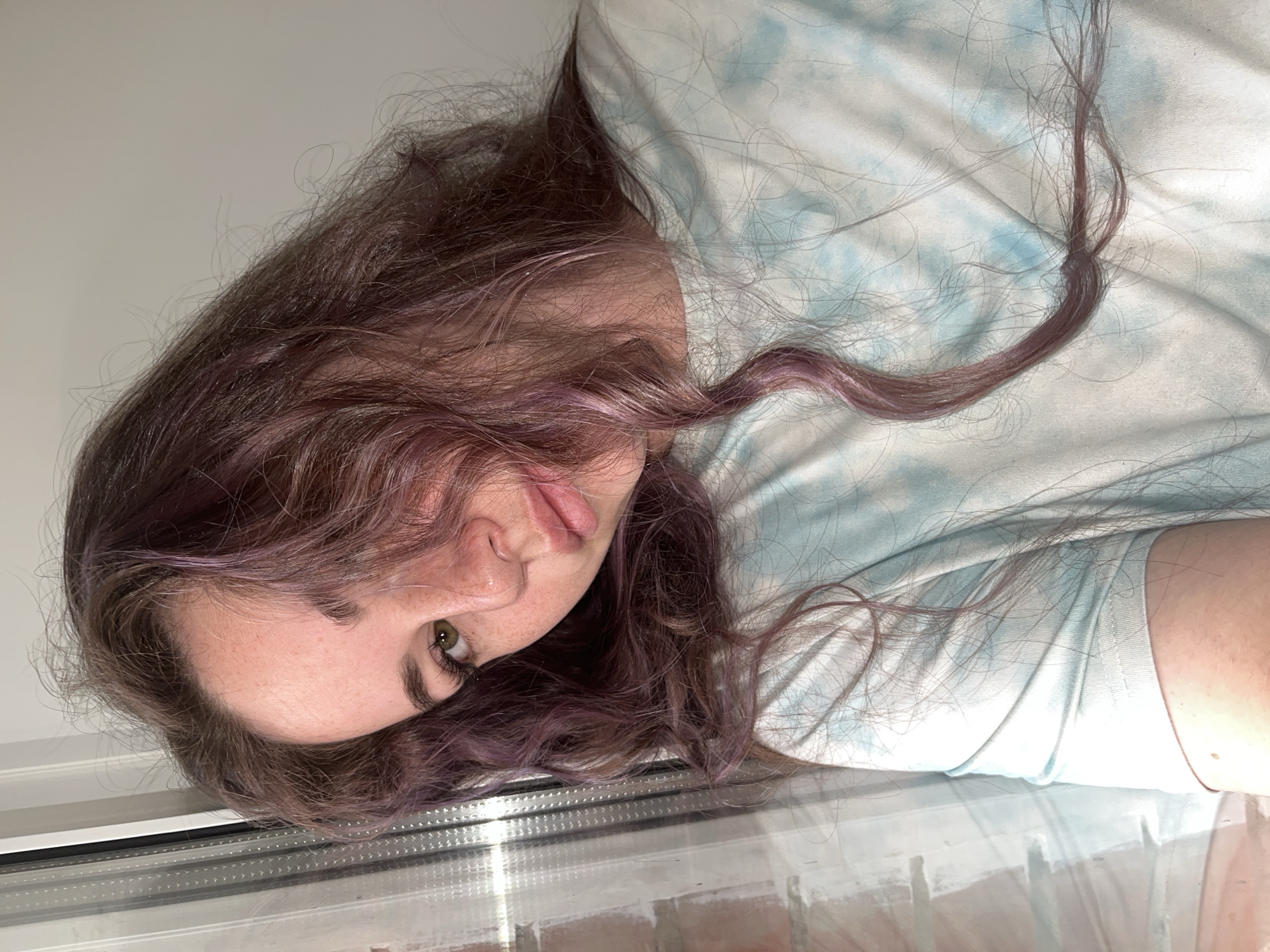 A woman with long, dark brown hair, some strands dyed a purplish-pink, is lying down, possibly on a bed or a flat surface. She is wearing a light blue tie-dye t-shirt. Her face is visible, with one eye clearly seen. The lower portion of the image shows a reflection in what appears to be a window or a smooth, reflective surface. The background suggests an indoor setting. There is no indication of location in Chaikovsky, Russia or any specific geographic location.