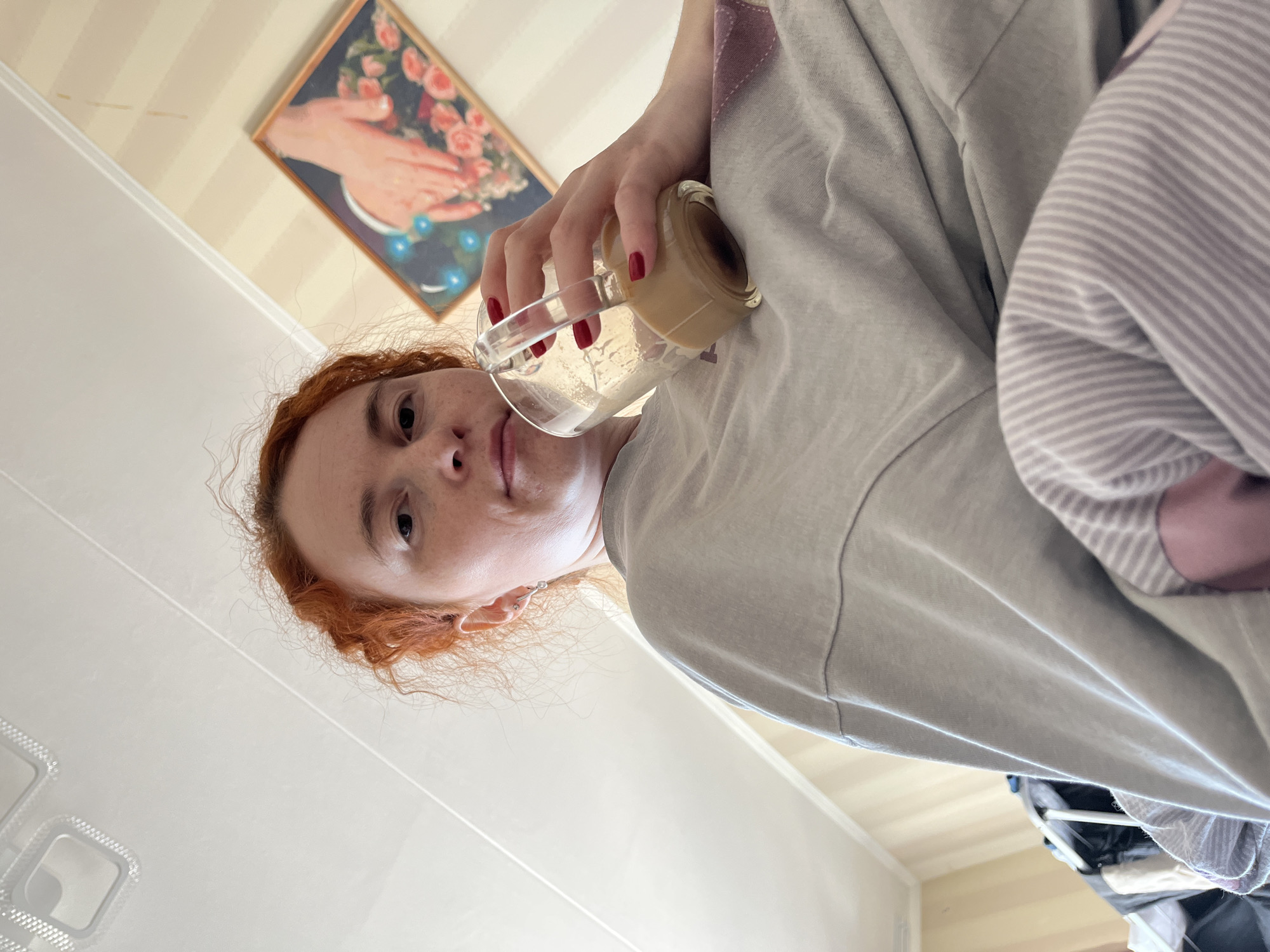 A woman with reddish-orange hair and red nail polish is tilting a clear glass mug, partially filled with a light-colored liquid, towards the camera. She is wearing a light grey T-shirt and a striped shirt is visible underneath. The setting appears to be indoors, possibly in an apartment in Chaikovsky, Russia, based on the background. A painting hangs on the wall with cream and beige striped wallpaper. White cabinets or storage units are visible in the lower left of the frame. The photograph is taken from a low angle, looking up at the woman.
